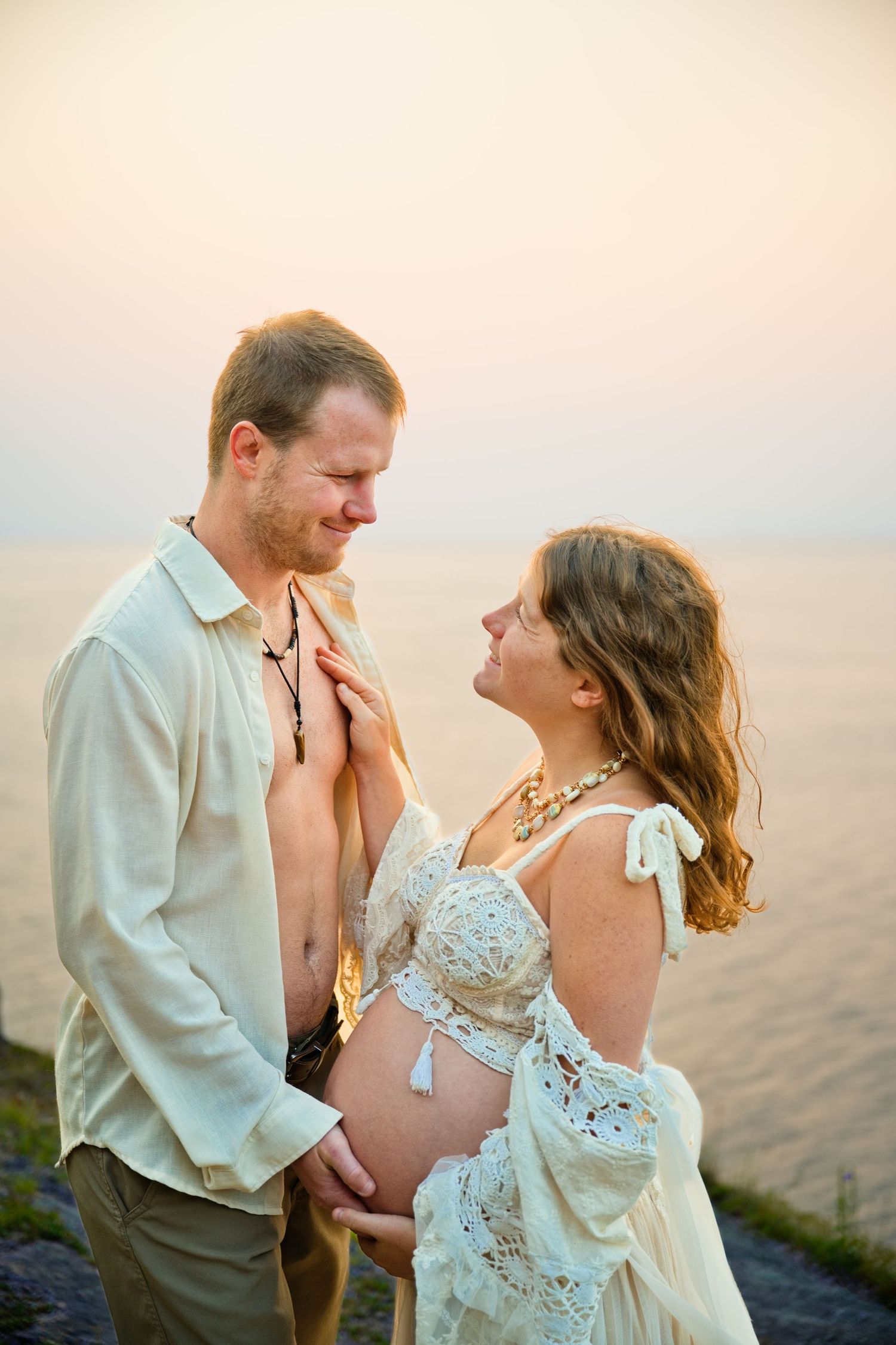 Pregnant couple sharing a tender moment at sunset by the sea, woman in lace dress cradling her baby bump.