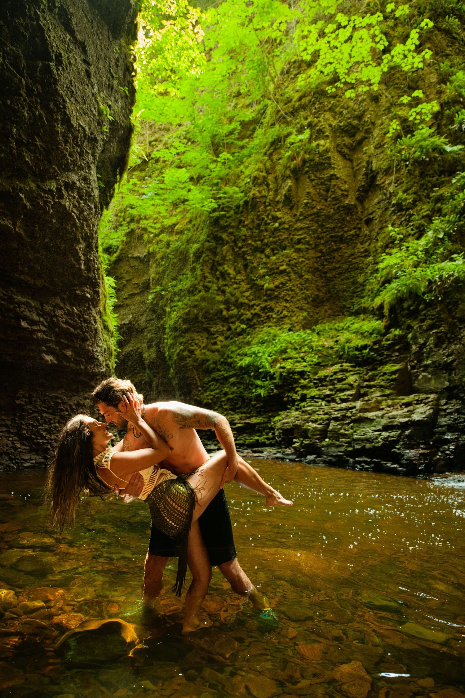 Couple sharing a romantic kiss in a lush green canyon with rocky walls and a shallow stream.