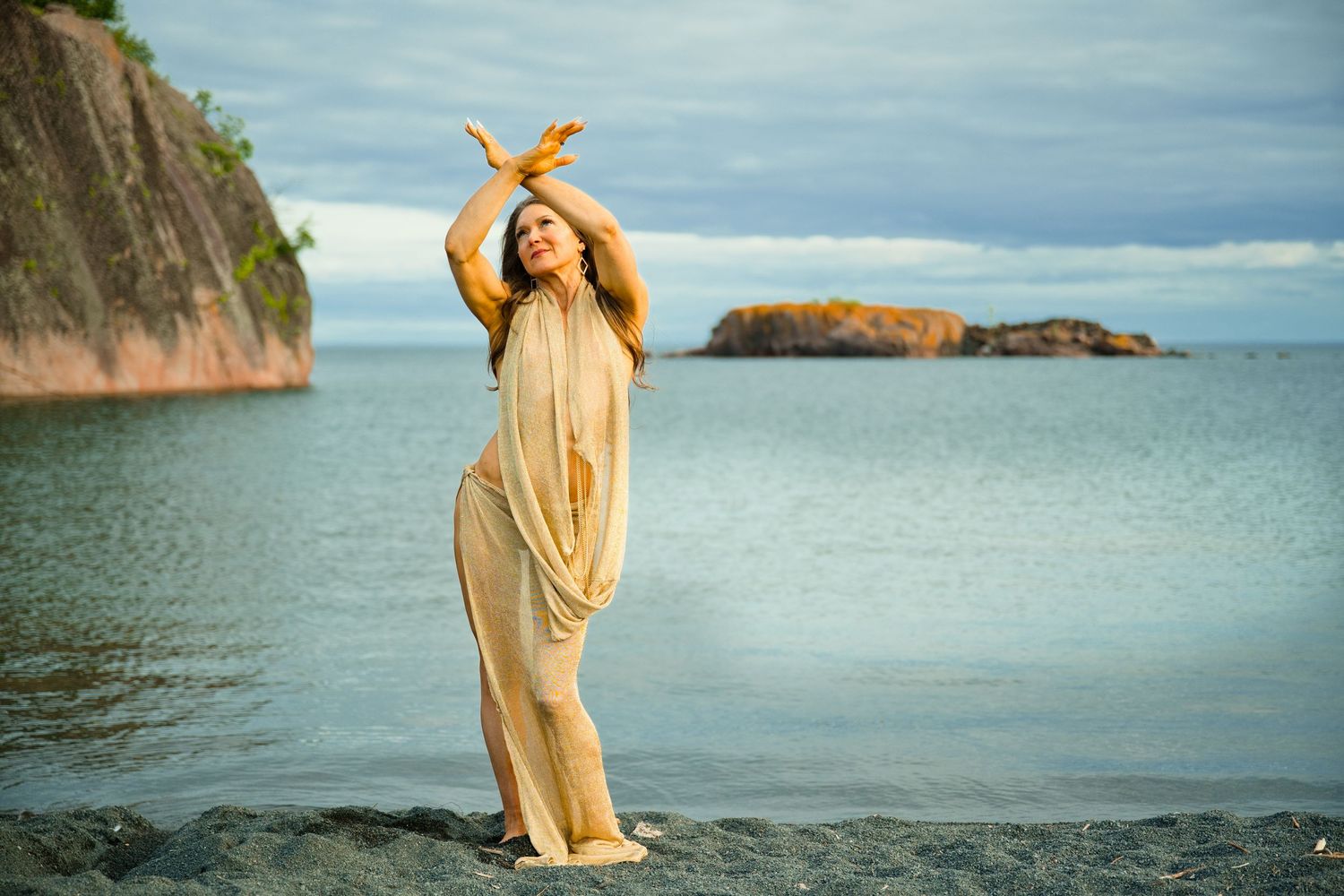 Woman in flowing gold dress poses gracefully on rocky shore with dramatic cliffs and ocean in background.