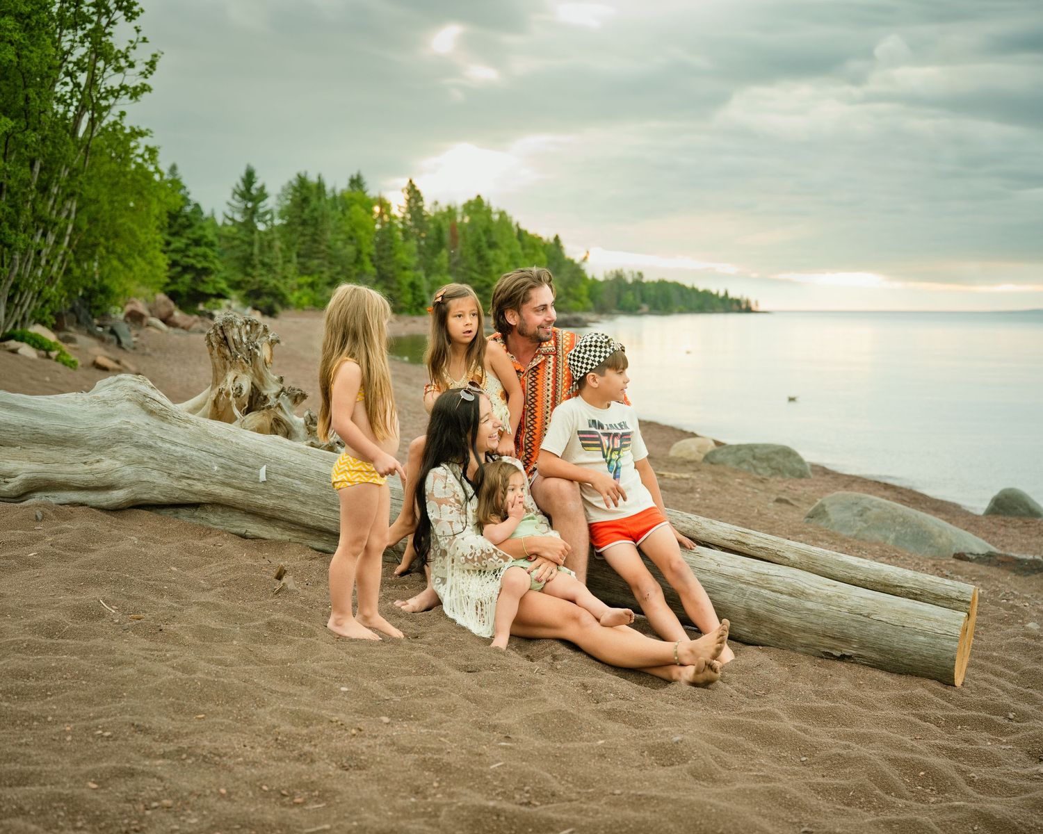 Family with four children sitting on driftwood log on sandy beach with lake and forest backdrop at dusk.