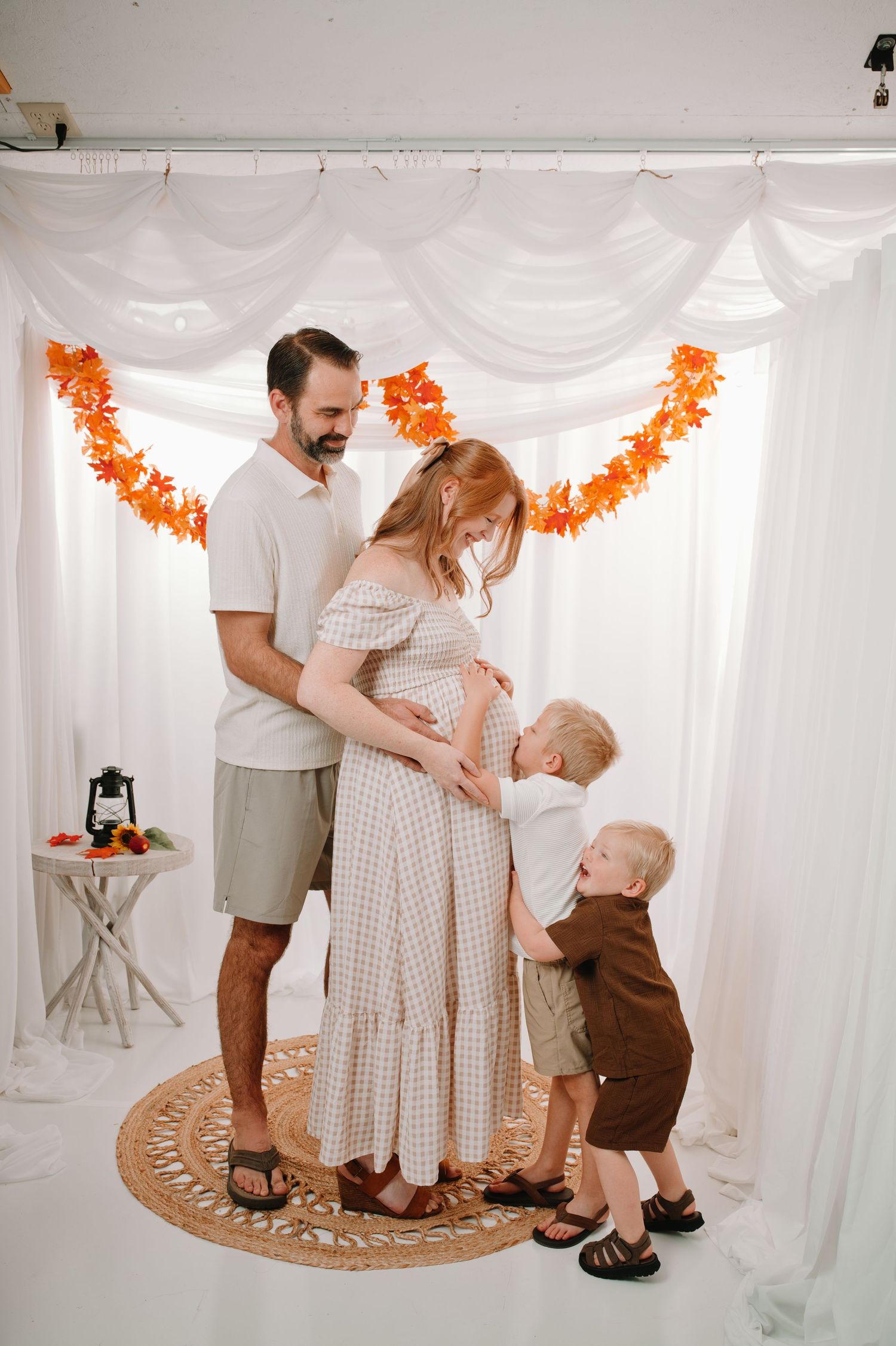 Family maternity photo with pregnant woman, husband, and two toddlers in white studio with fall leaf garland.