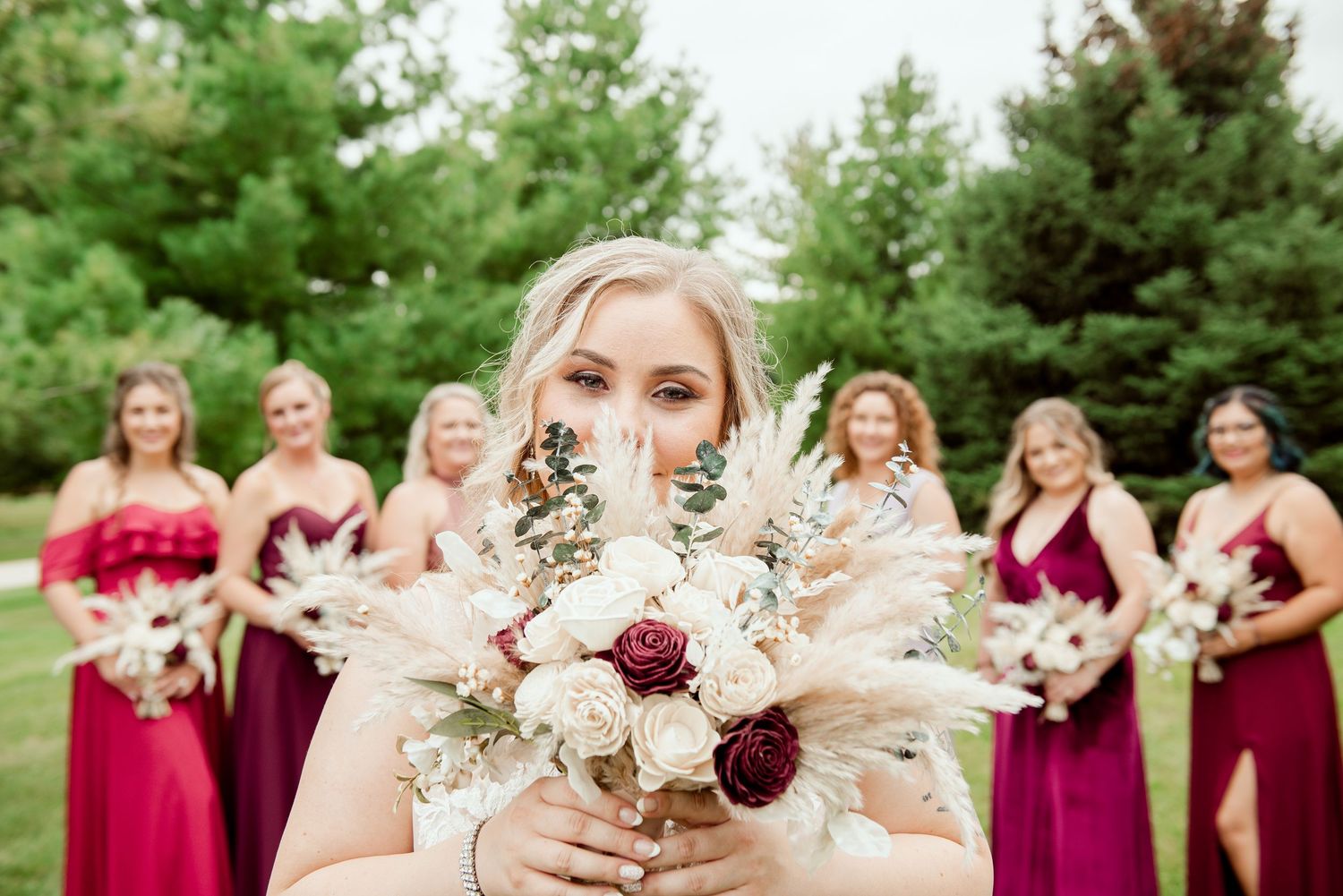 Bride holds romantic bouquet with burgundy and white flowers while bridesmaids stand in background.