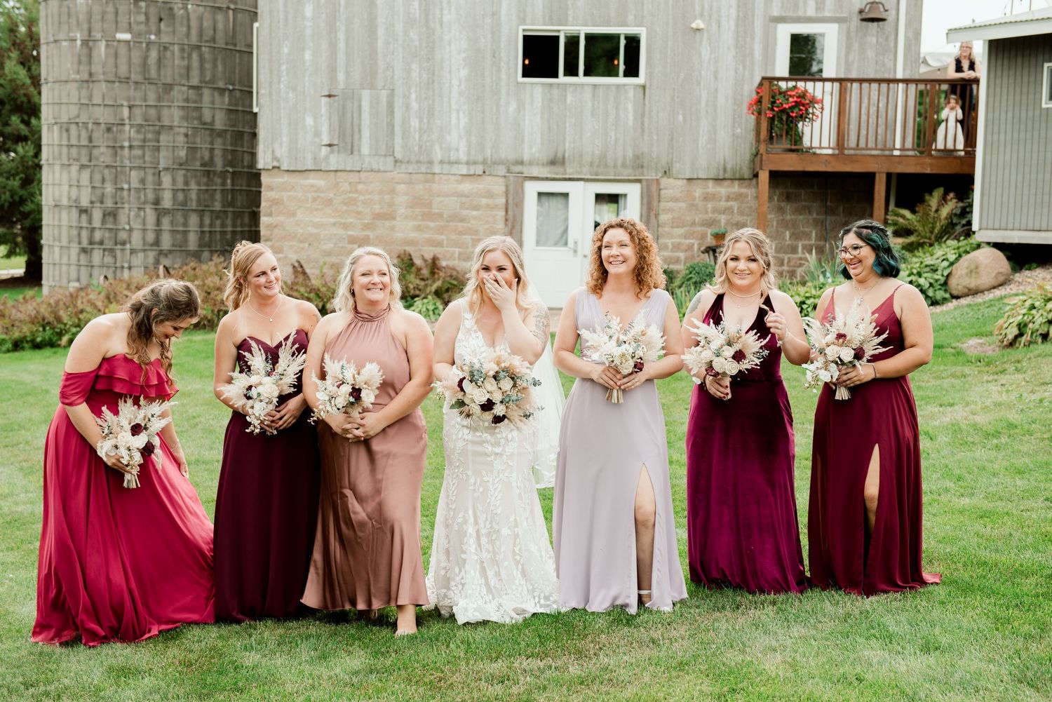 Bridal party poses in front of rustic barn wearing burgundy and blush tones.