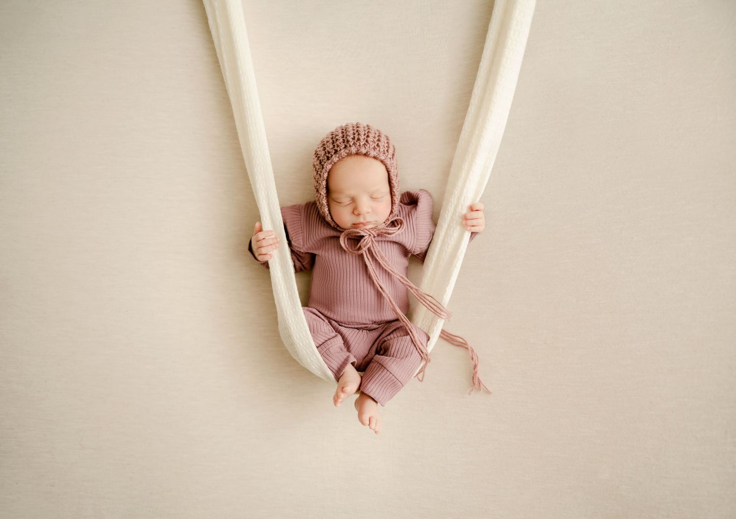 St Cloud newborn Photos of Newborn baby sleeping peacefully in a cream fabric hammock wearing a pink knit bonnet and matching outfit.