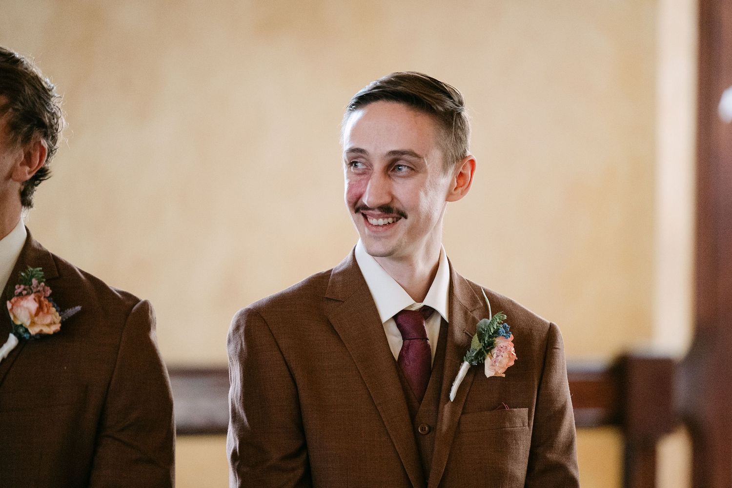 Central MN Wedding Photos | Smiling groom in brown suit with floral boutonniere at wedding ceremony.