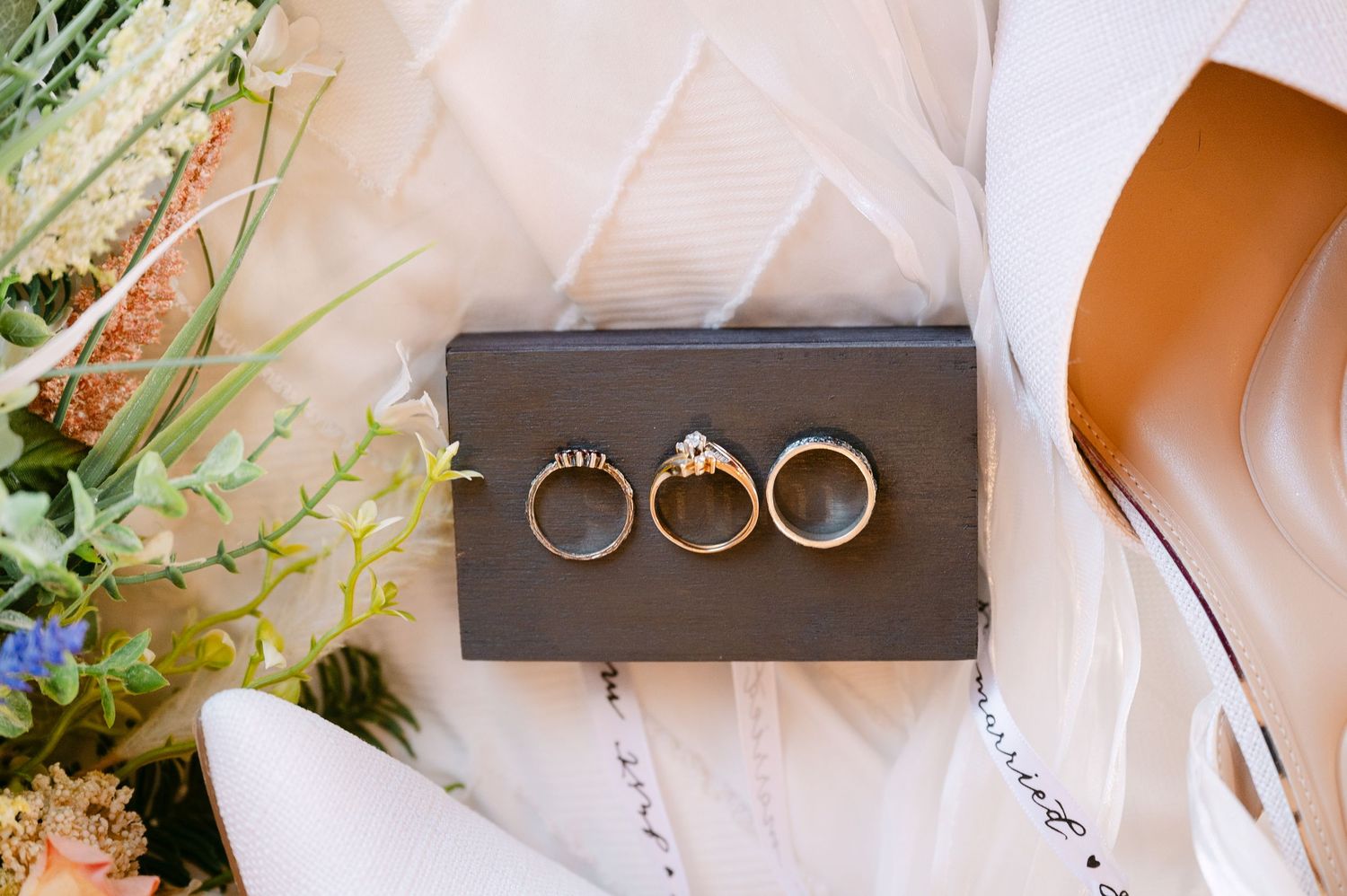 Central MN Wedding Photos | Three gold wedding rings displayed on a dark box with bridal bouquet and white heels in the background.