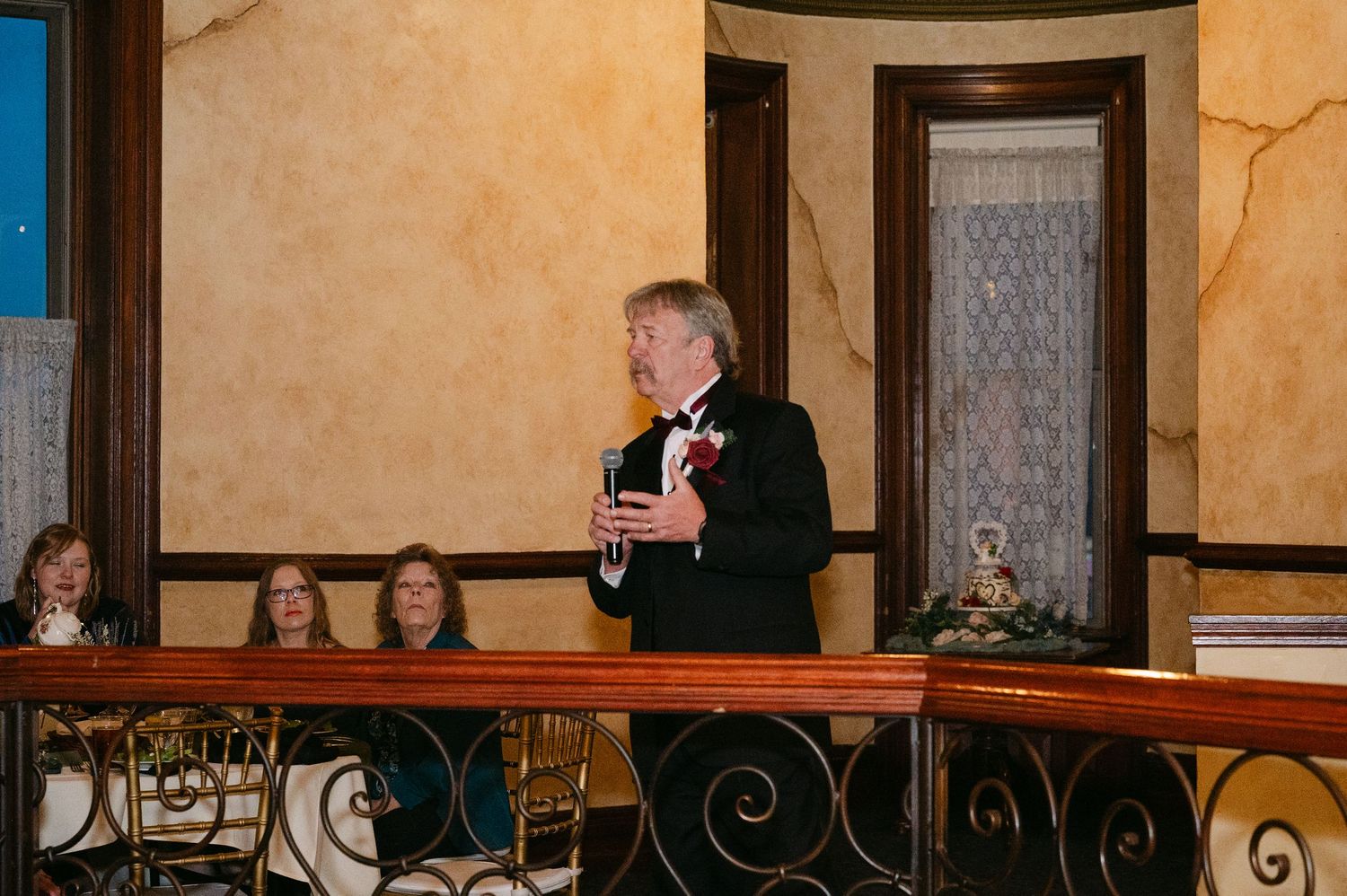 Central MN Wedding Photos | Man in tuxedo with boutonniere delivering speech at wedding reception on decorated balcony.