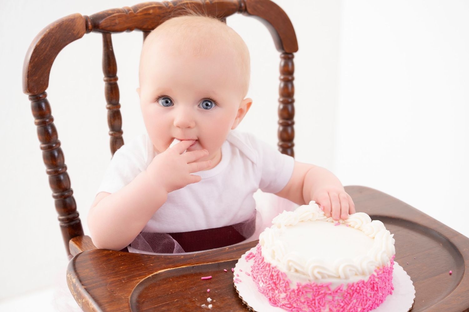 Central MN Cake Smash Photography - Cute baby with blue eyes sitting in wooden high chair, tasting pink and white birthday cake with finger.