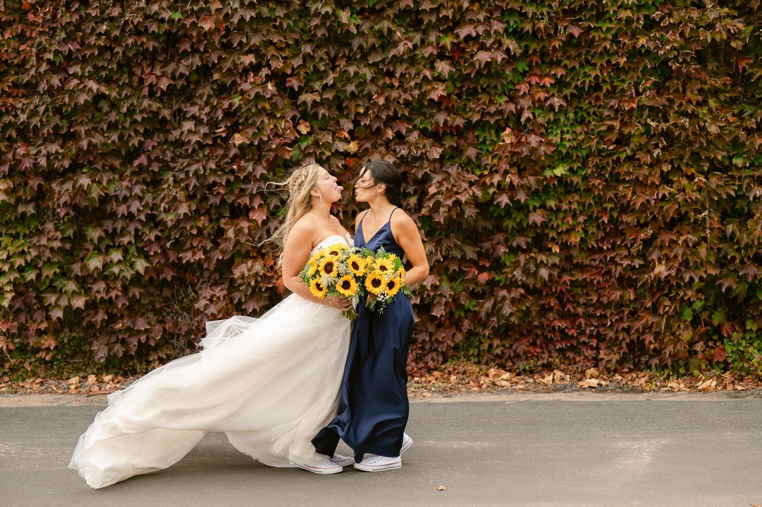 Central MN Wedding Photos | Bride in white gown and bridesmaid in navy dress holding sunflower bouquets laughing together outdoors.
