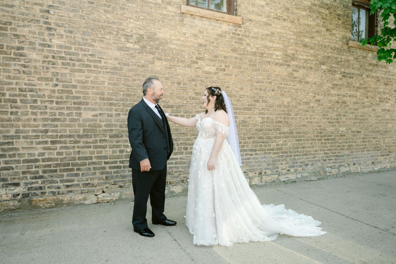Central MN Wedding Photos - Bride in lace off-shoulder gown shares first look moment with father in black suit against brick wall.