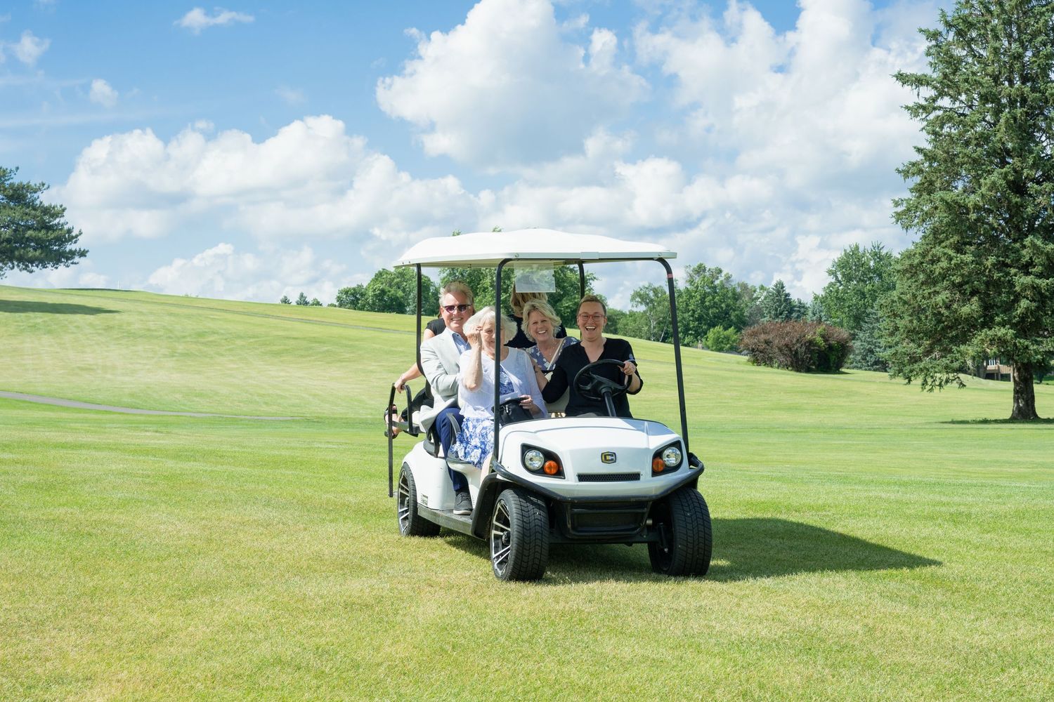 Three smiling guests riding white golf cart across lush green golf course under blue sky.