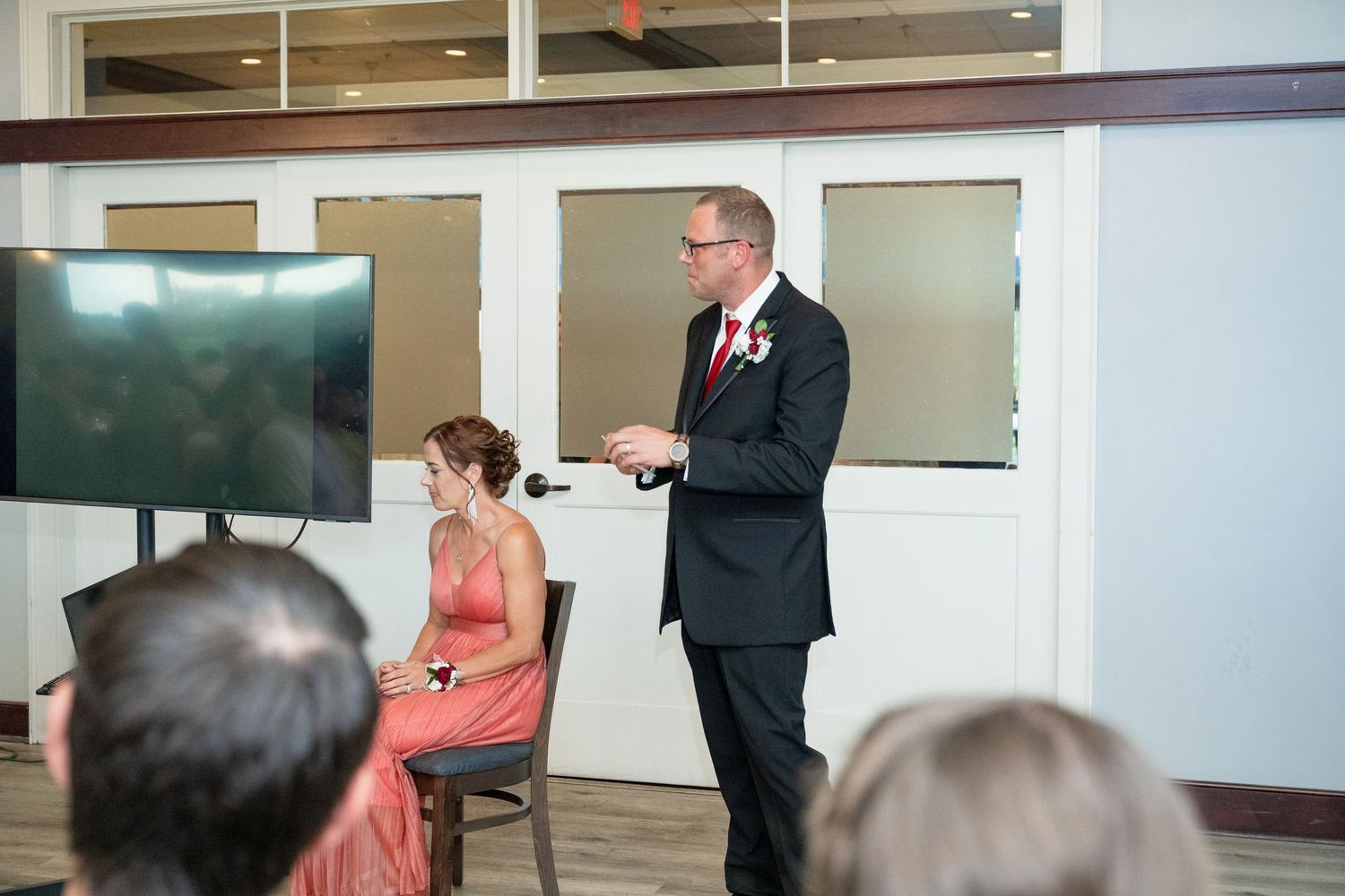 Groom in black suit giving a speech at wedding reception while bride sits in coral dress nearby.