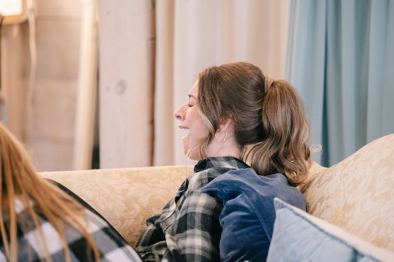crooked willow weddings A smiling woman with a ponytail relaxes on a beige couch wearing a plaid flannel shirt in a cozy room.
