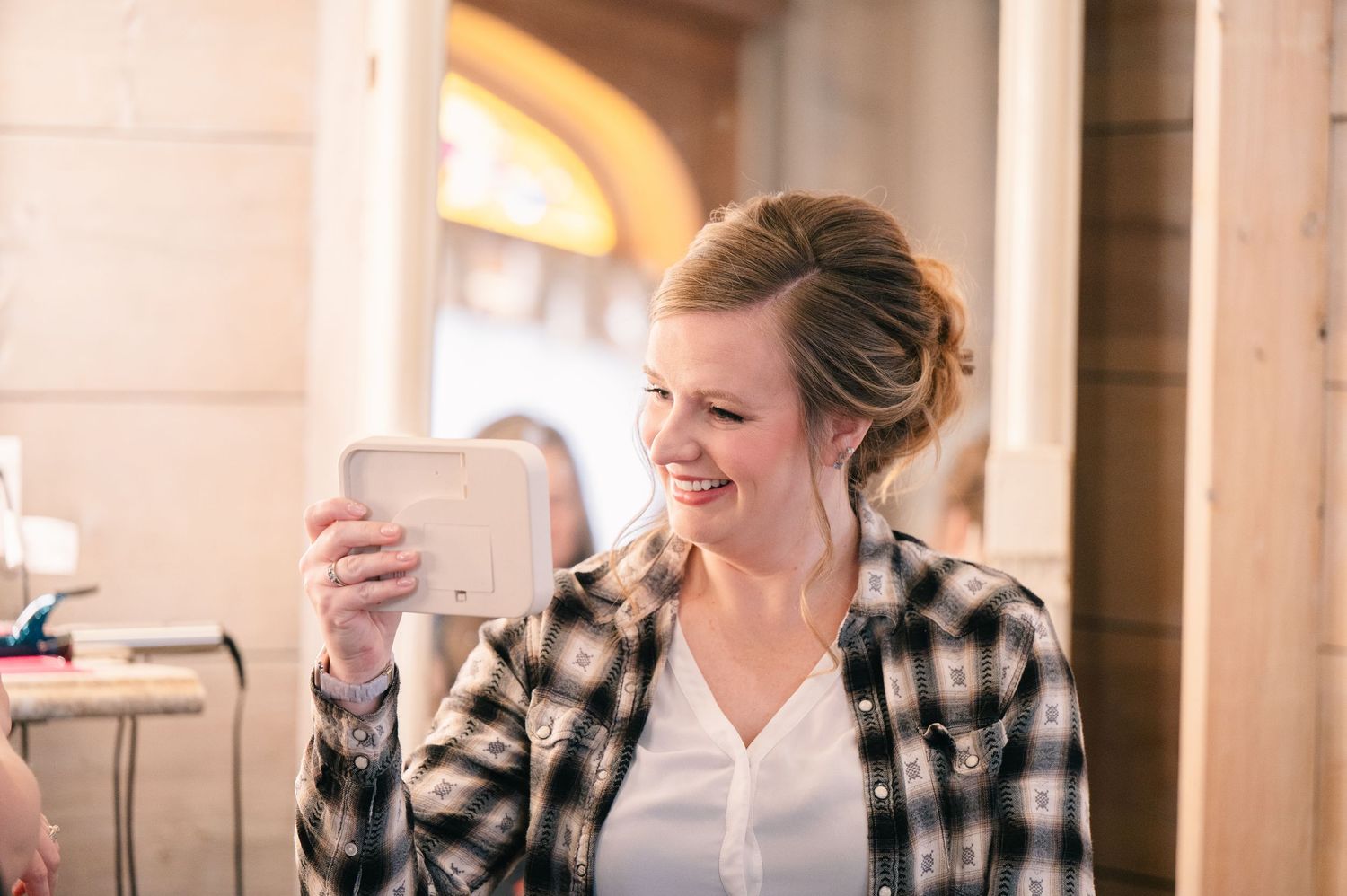 Smiling woman in plaid shirt holding a small mirror indoors at a rustic venue.