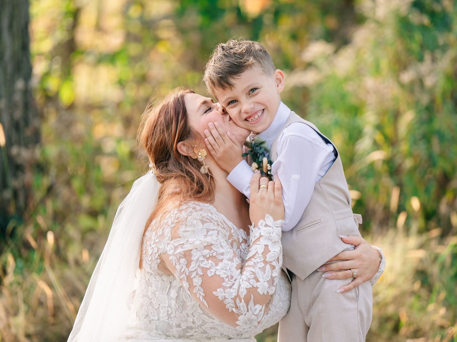 Bride in lace wedding gown kisses the cheek of a smiling young boy in a tan suit outdoors.