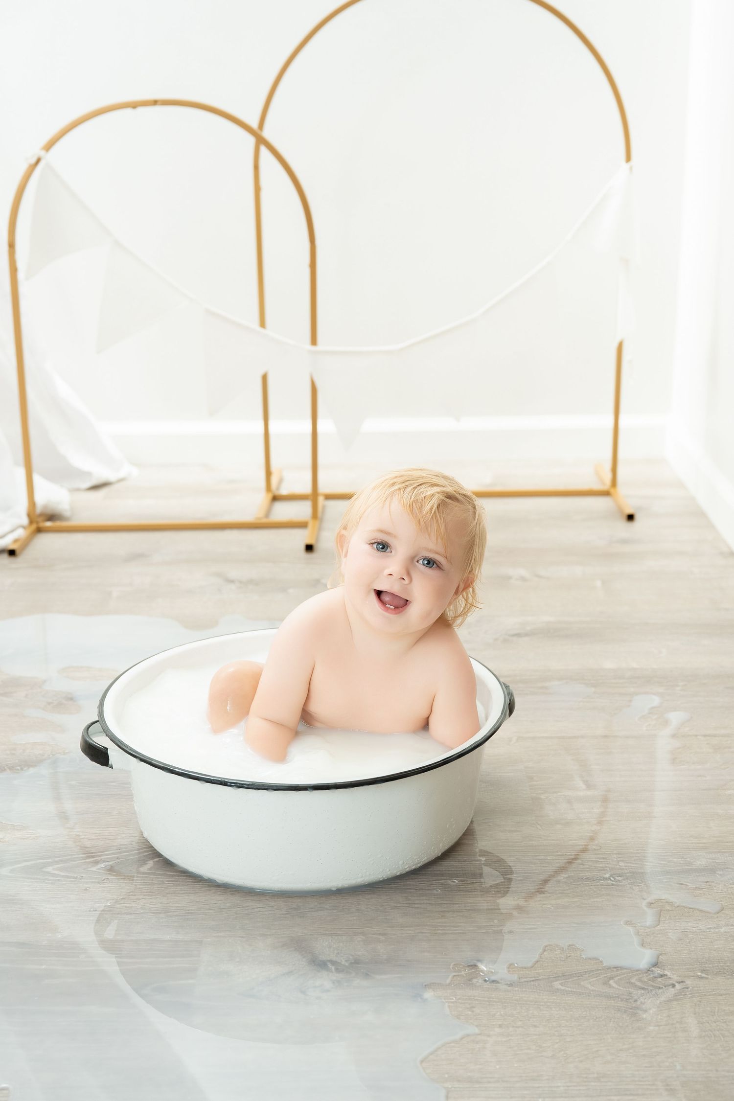 A small child sits in a white enamel basin during a photo session with gold arch decorations in the background. Princeton MN Milestone, Mate