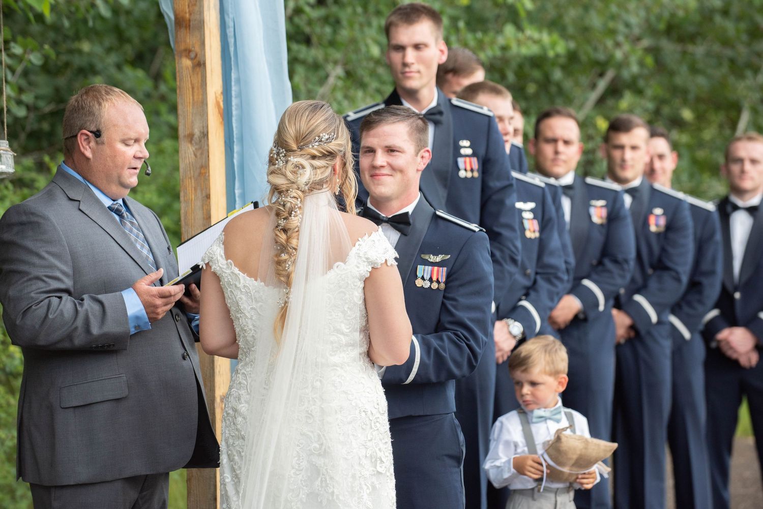 An outdoor wedding ceremony takes place under a wooden arch draped with white fabric. Zimmerman MN Weddings