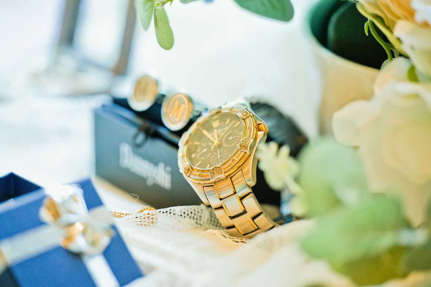 Gold chronograph watch with cufflinks and Tiffany blue jewelry box on a decorated table with white flowers.