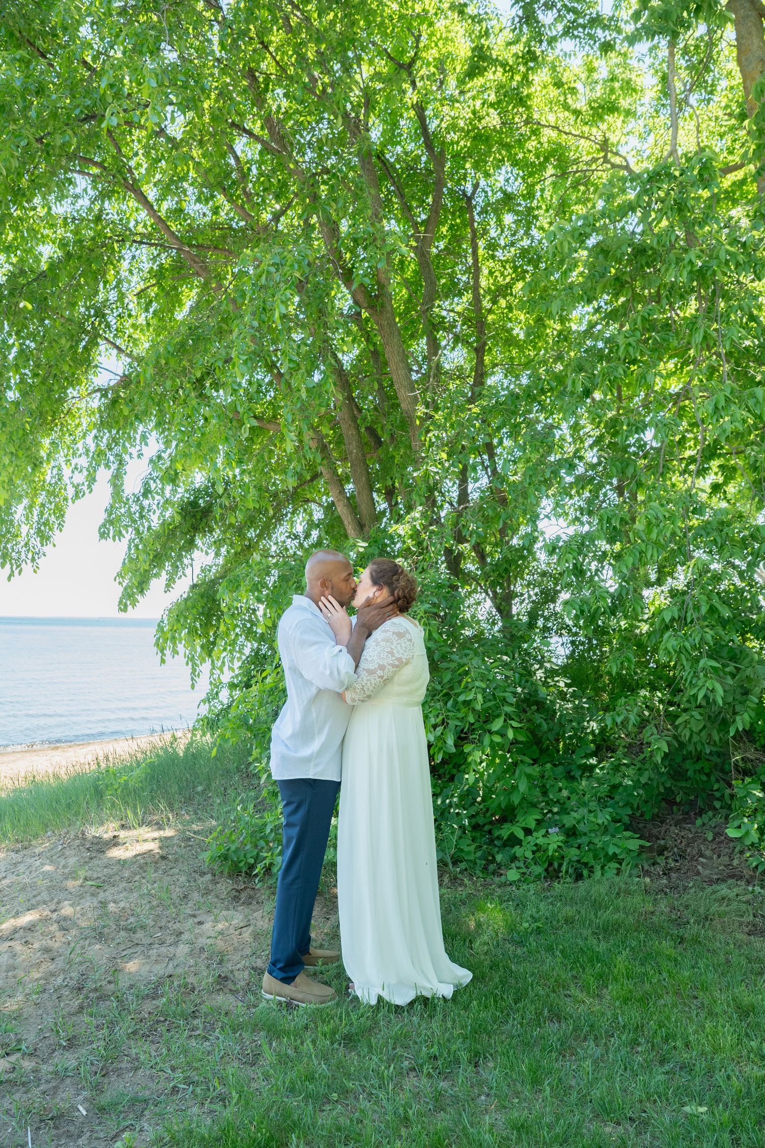 Bride and groom sharing a kiss under a large green willow tree near a lakeside beach.