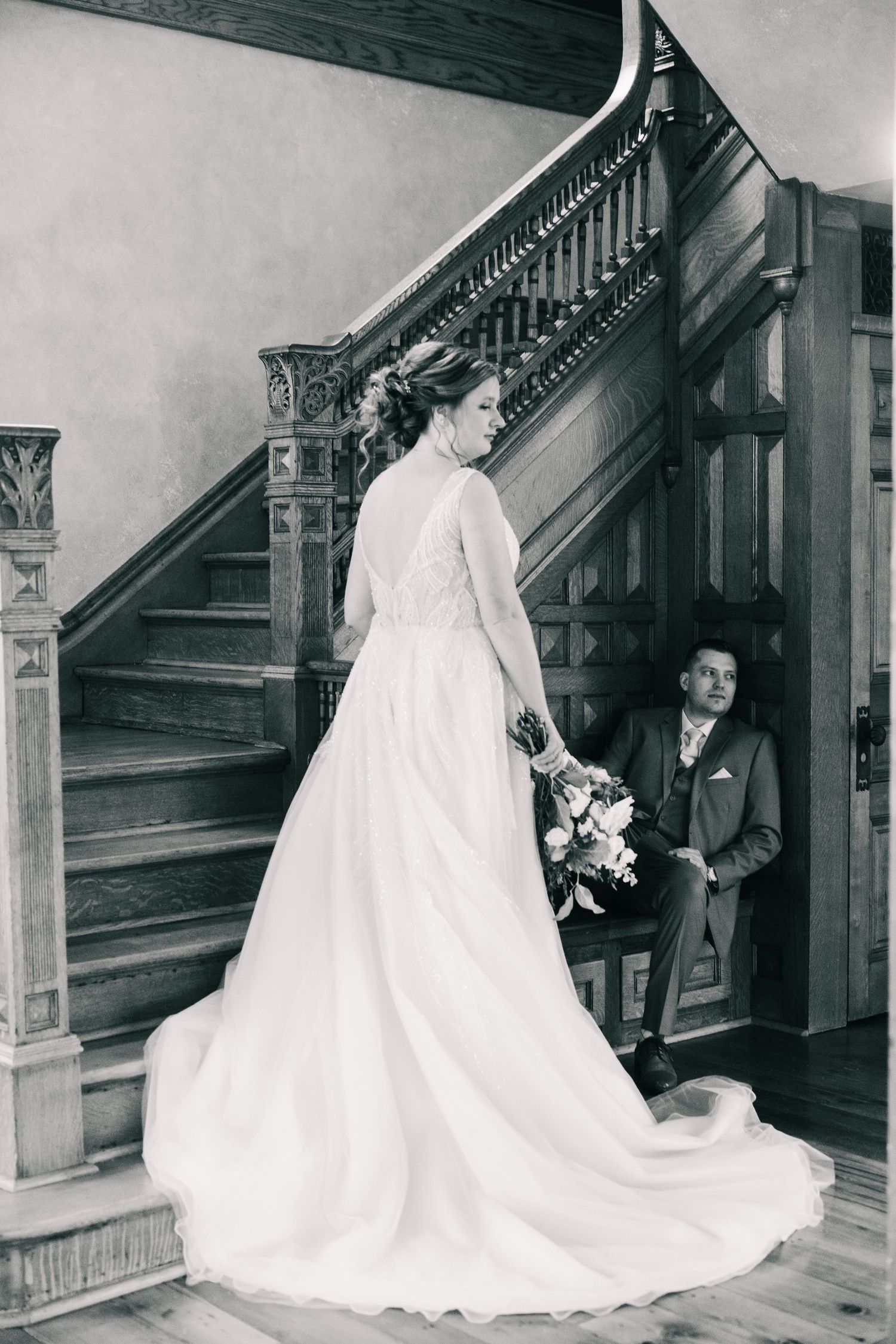 Black and white photo of bride in flowing gown with groom seated on ornate wooden staircase.