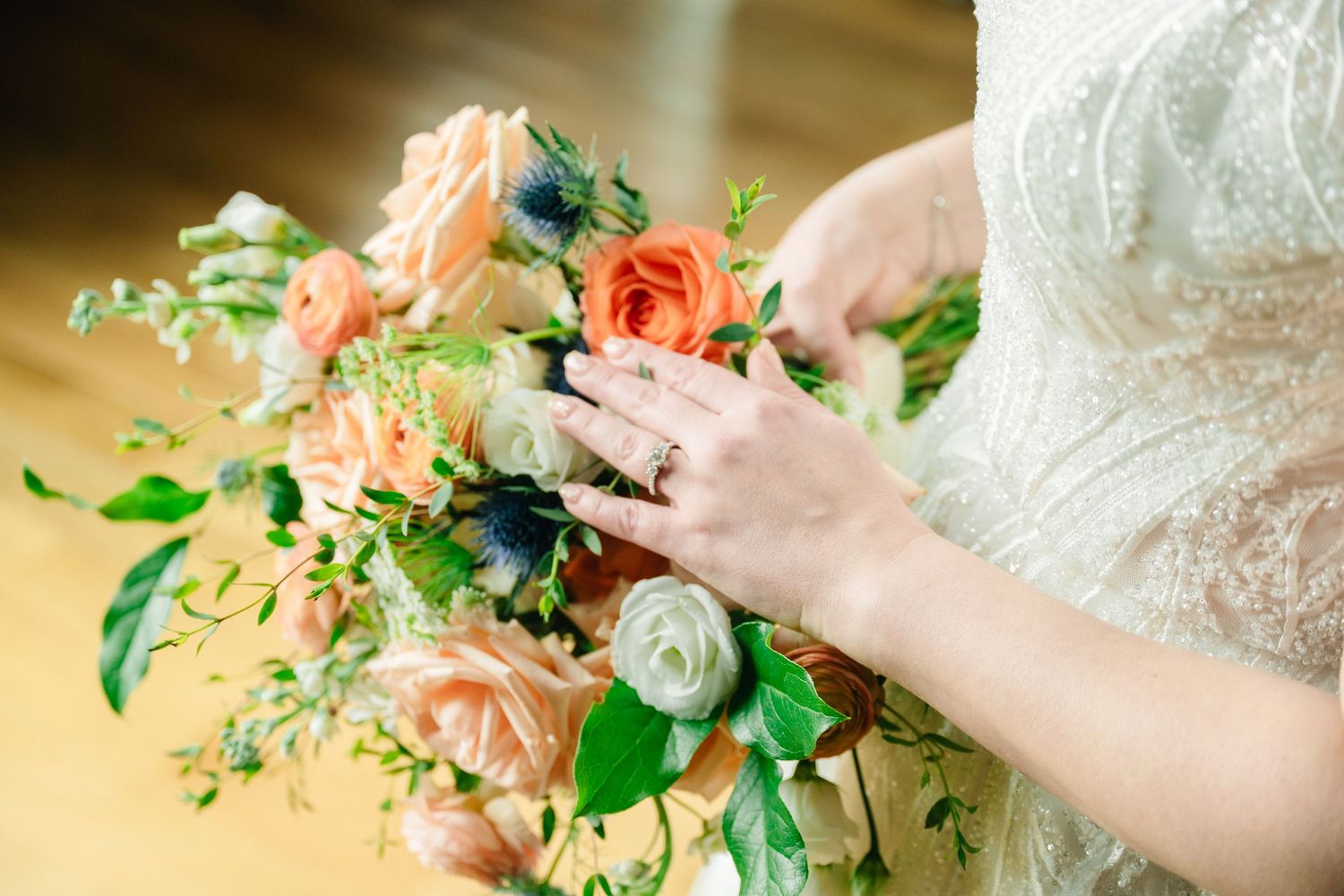 Close-up of bride holding a lush peach, white, and blue floral bouquet with greenery, wearing diamond ring.