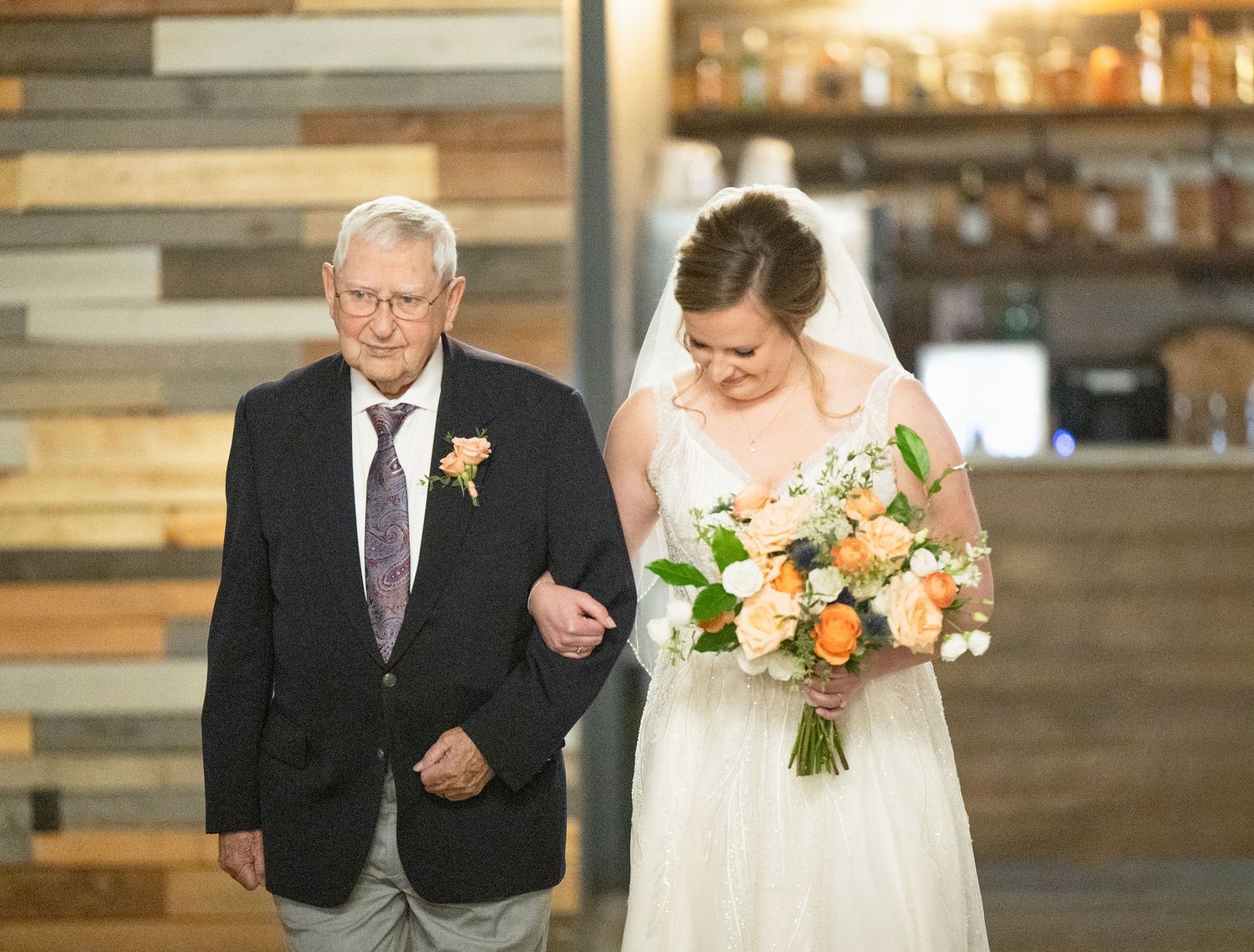 Bride in white dress walks down aisle with elderly father, holding peach and orange bouquet.