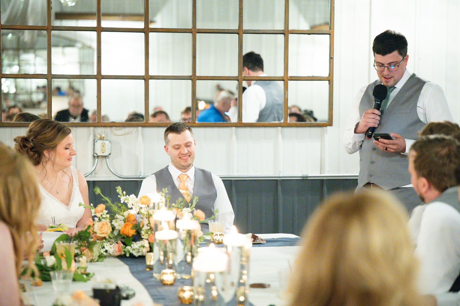 Best man gives a heartfelt wedding toast as bride and groom smile at decorated reception table with candles.