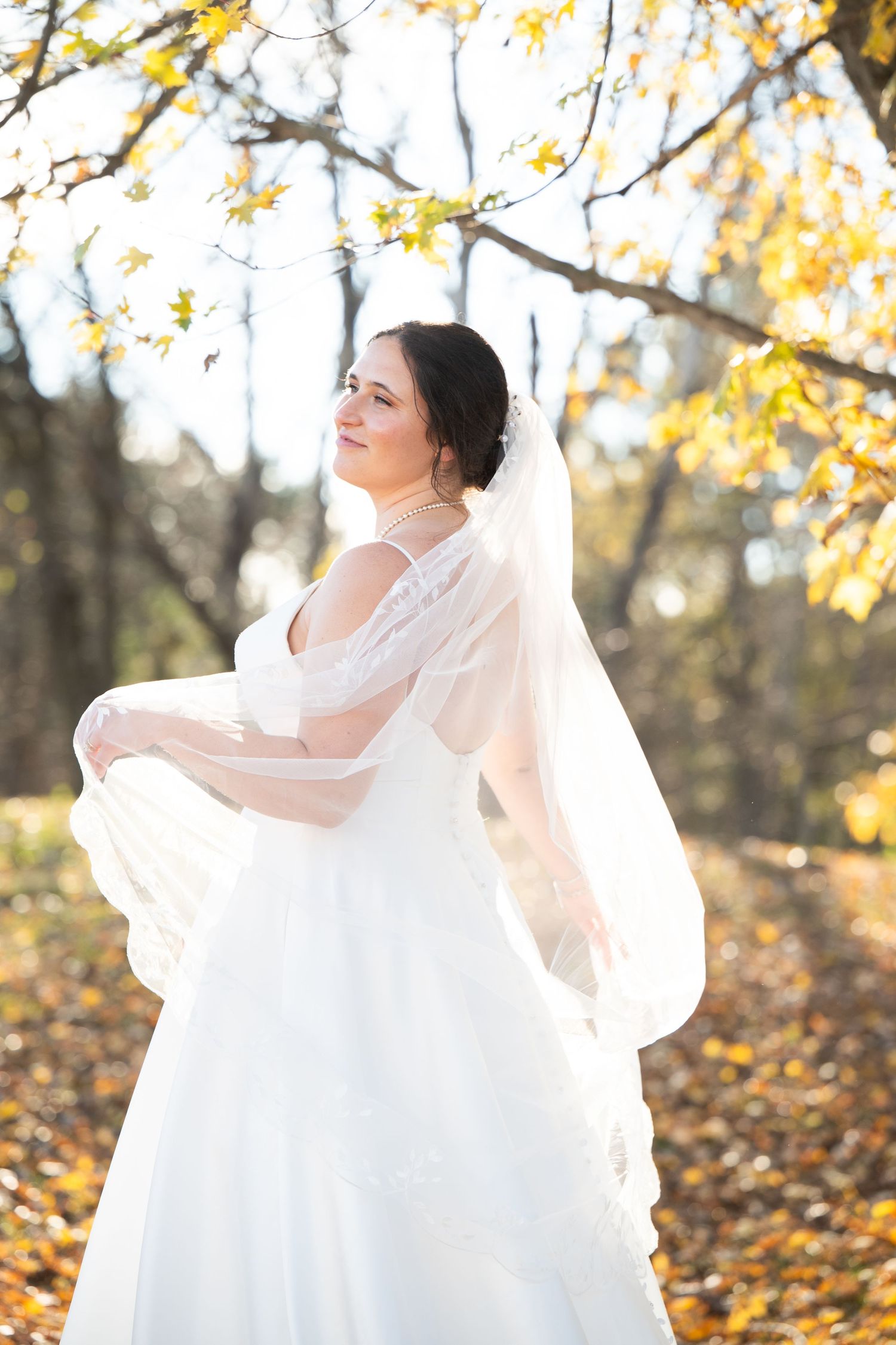 A bride in a flowing white dress twirls in autumn sunlight among fallen leaves.