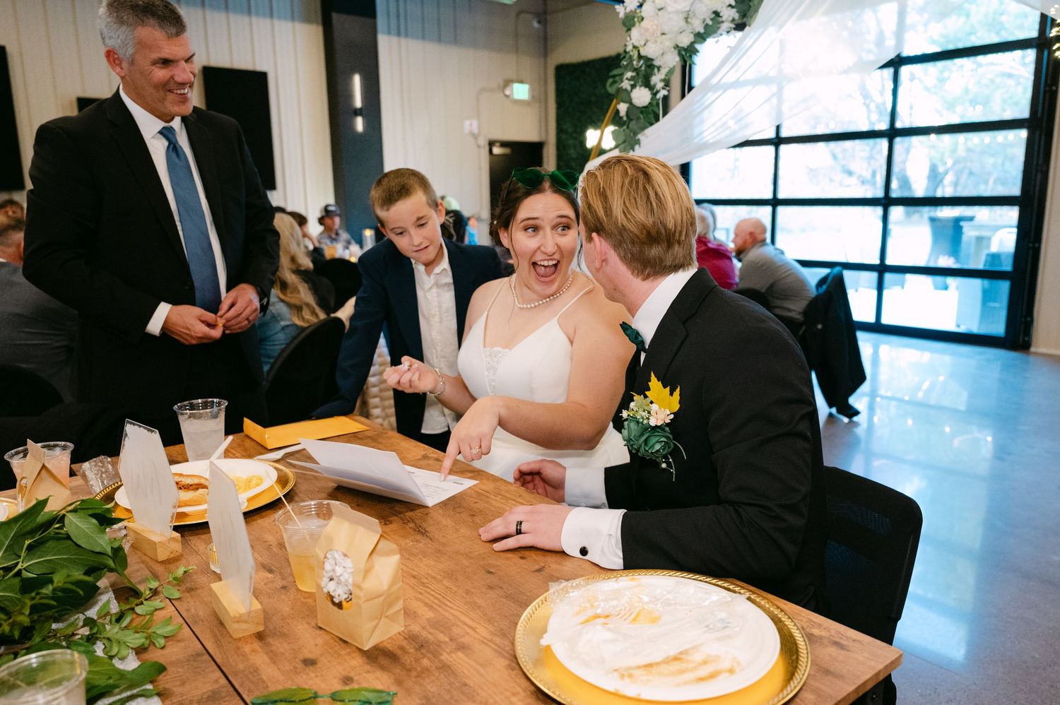 Wedding party gathers around wooden tables during an elegant indoor reception celebration.