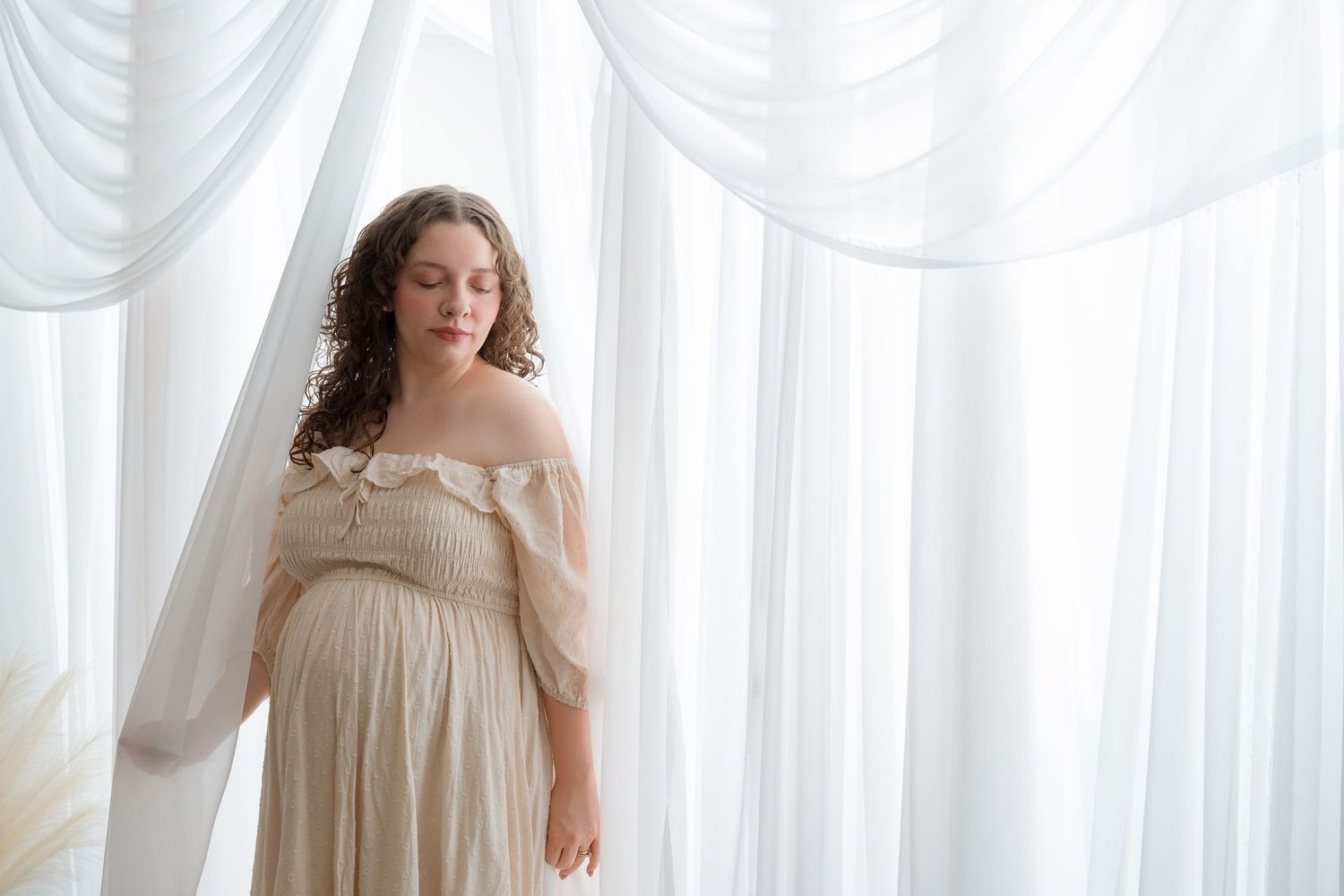 Woman in flowing cream-colored dress poses elegantly against white curtained backdrop in ethereal boudoir setting.