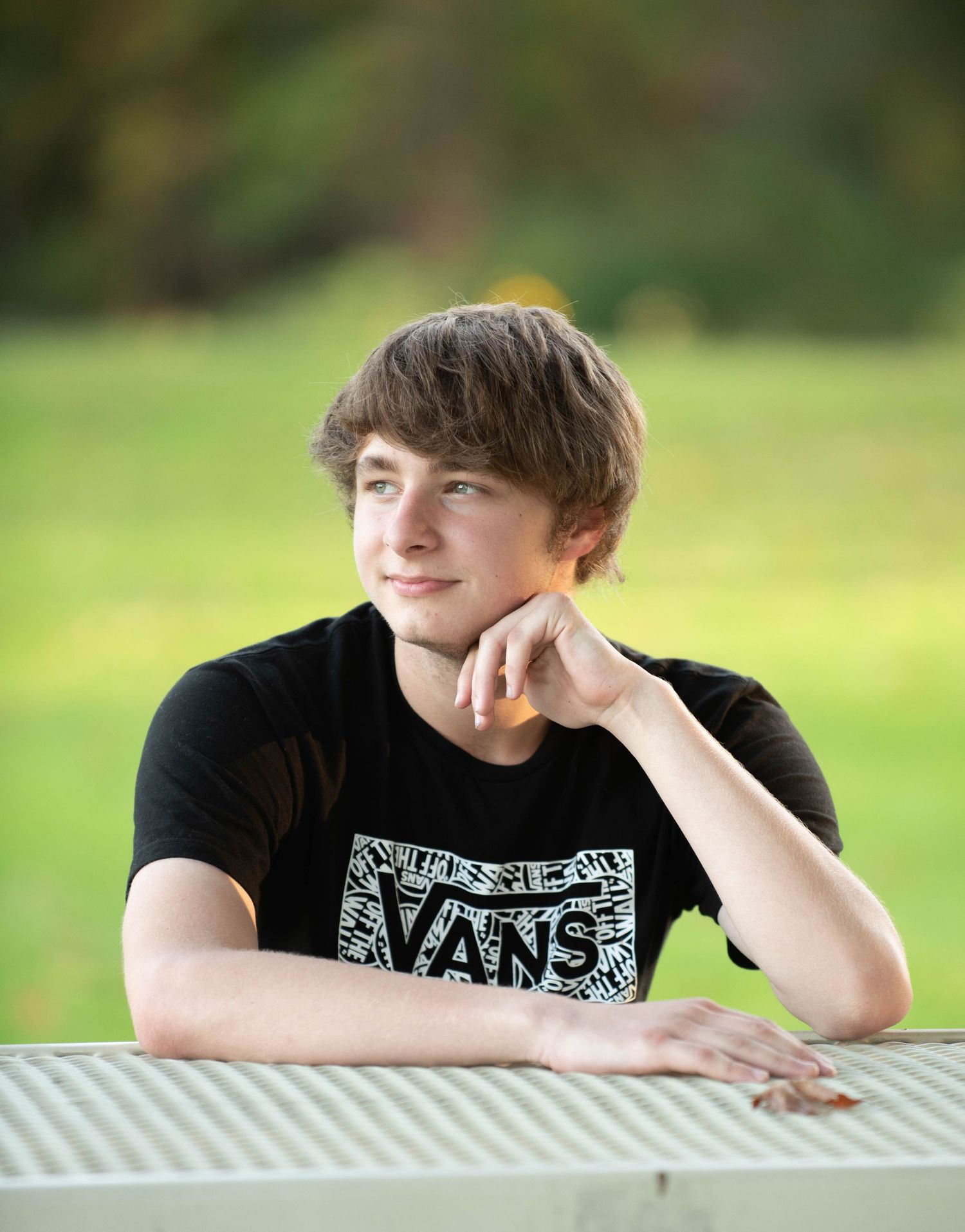 Person in black Vans t-shirt leaning on white surface with green outdoor backdrop.
