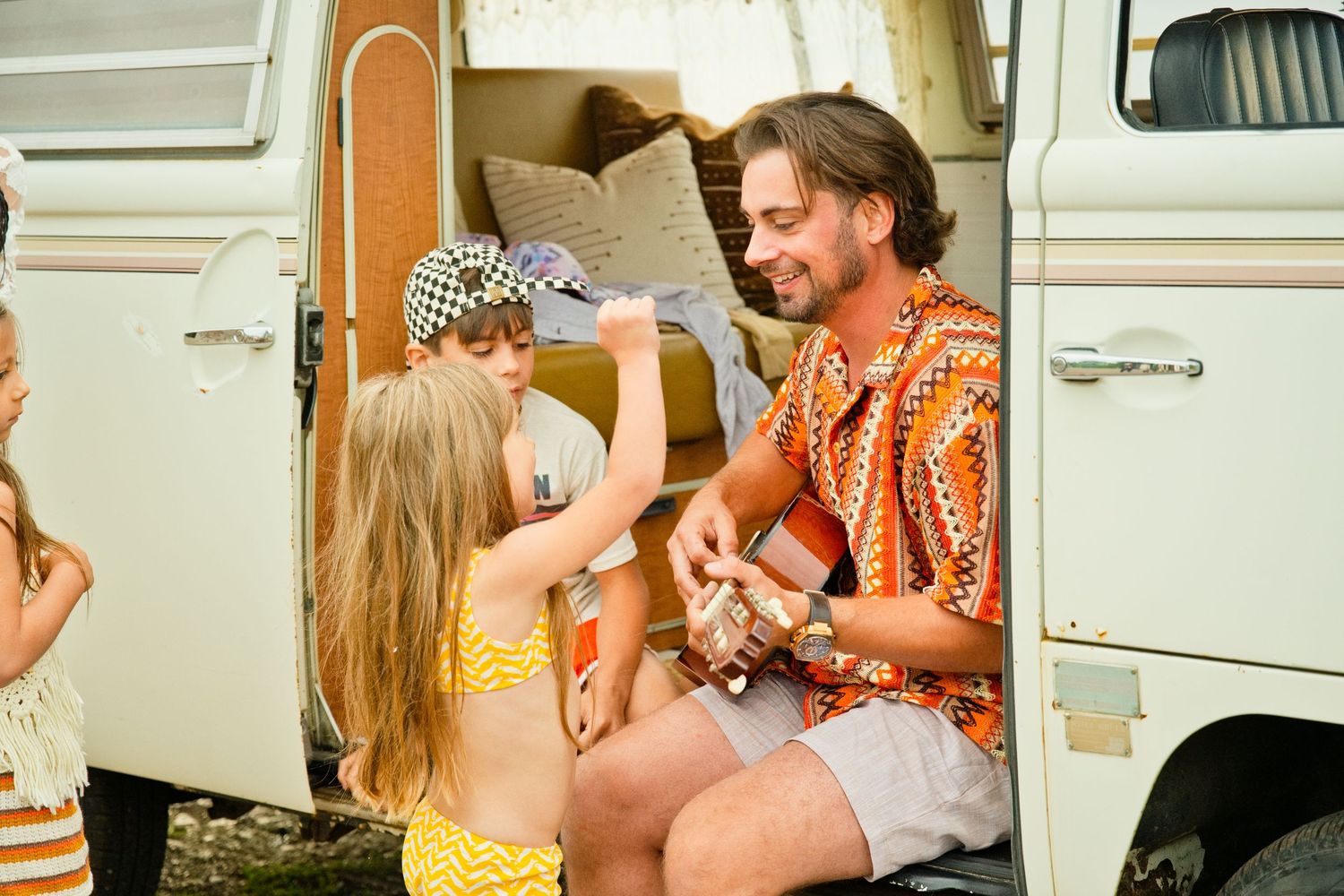 Man playing guitar for children beside a vintage white camper van on a sunny summer day.