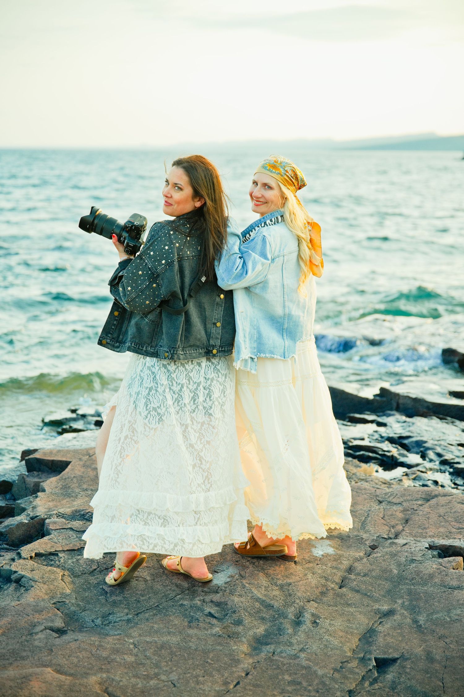 Two women in white lace dresses pose on rocky shore, one holding camera, ocean waves behind them.