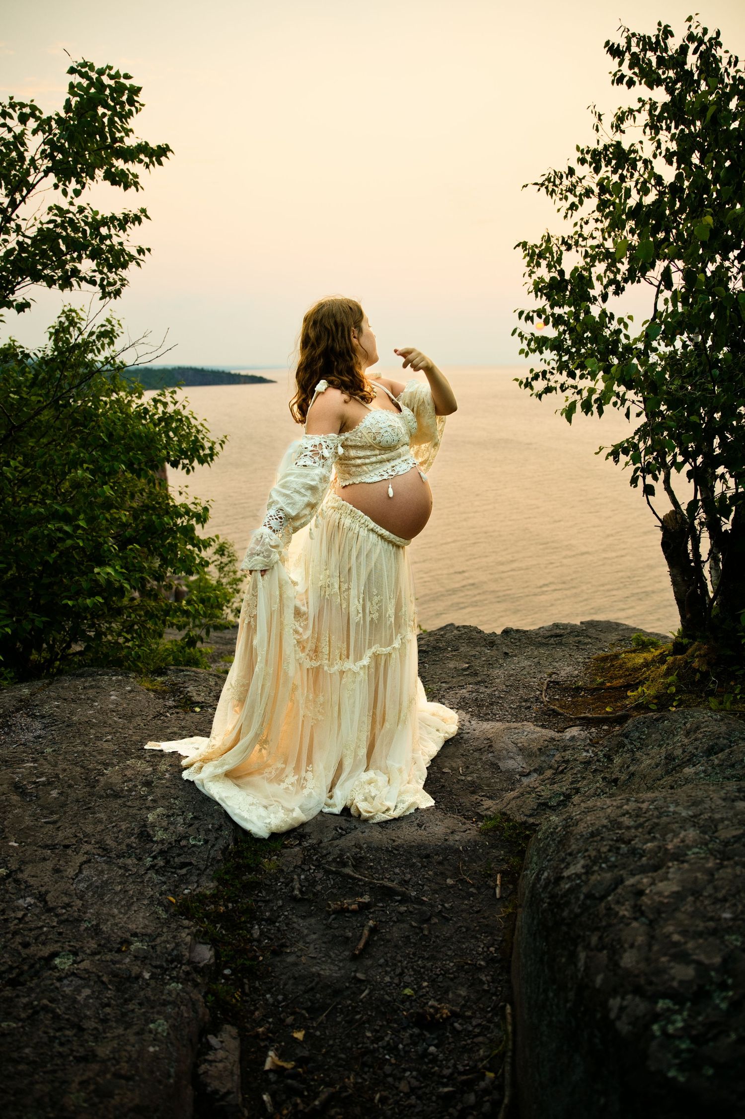 Pregnant woman in elegant lace gown standing on cliff overlooking serene ocean at sunset.