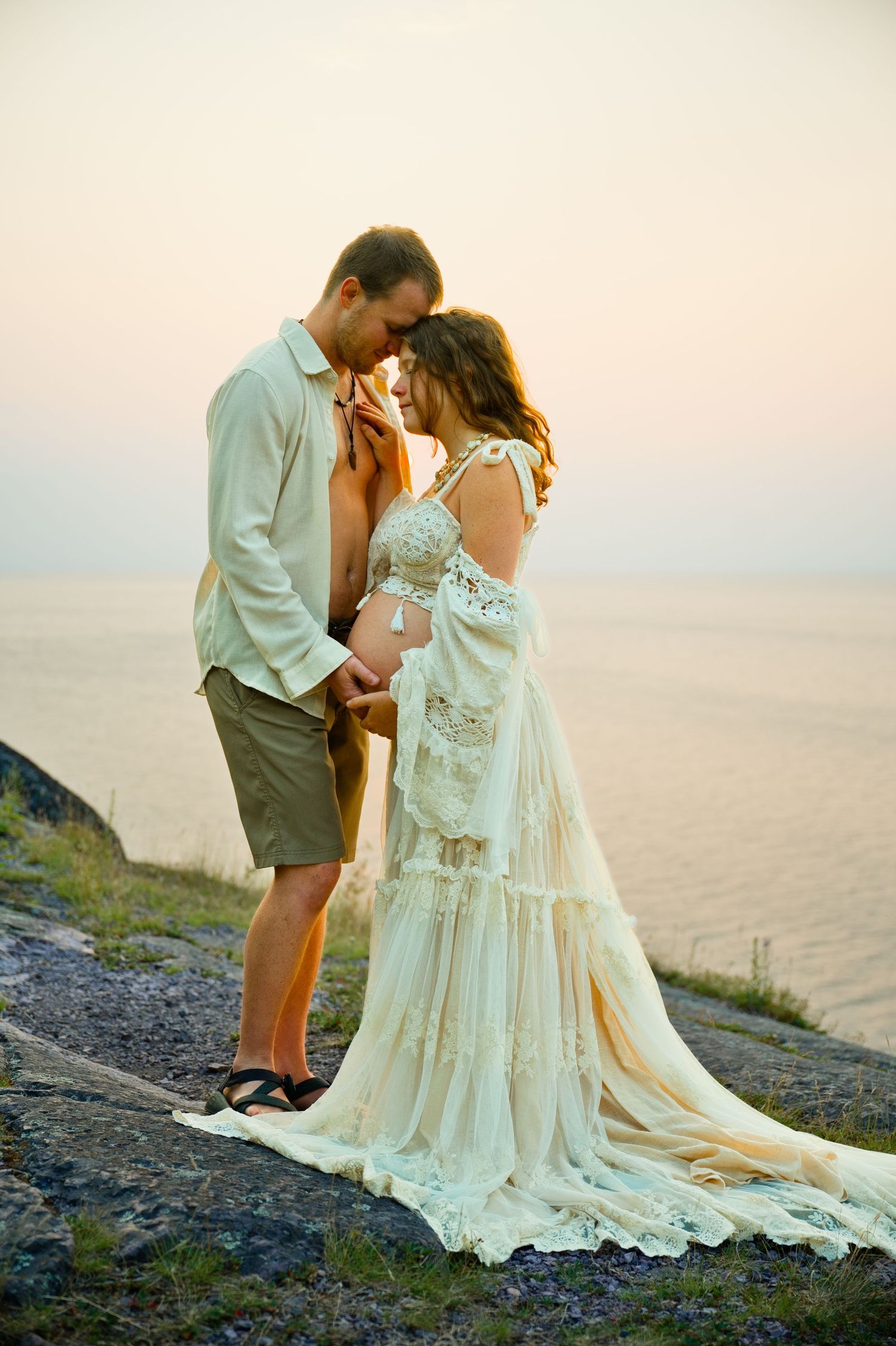 Couple in romantic embrace on rocky cliff at sunset, woman in elegant boho lace wedding dress, ocean backdrop.