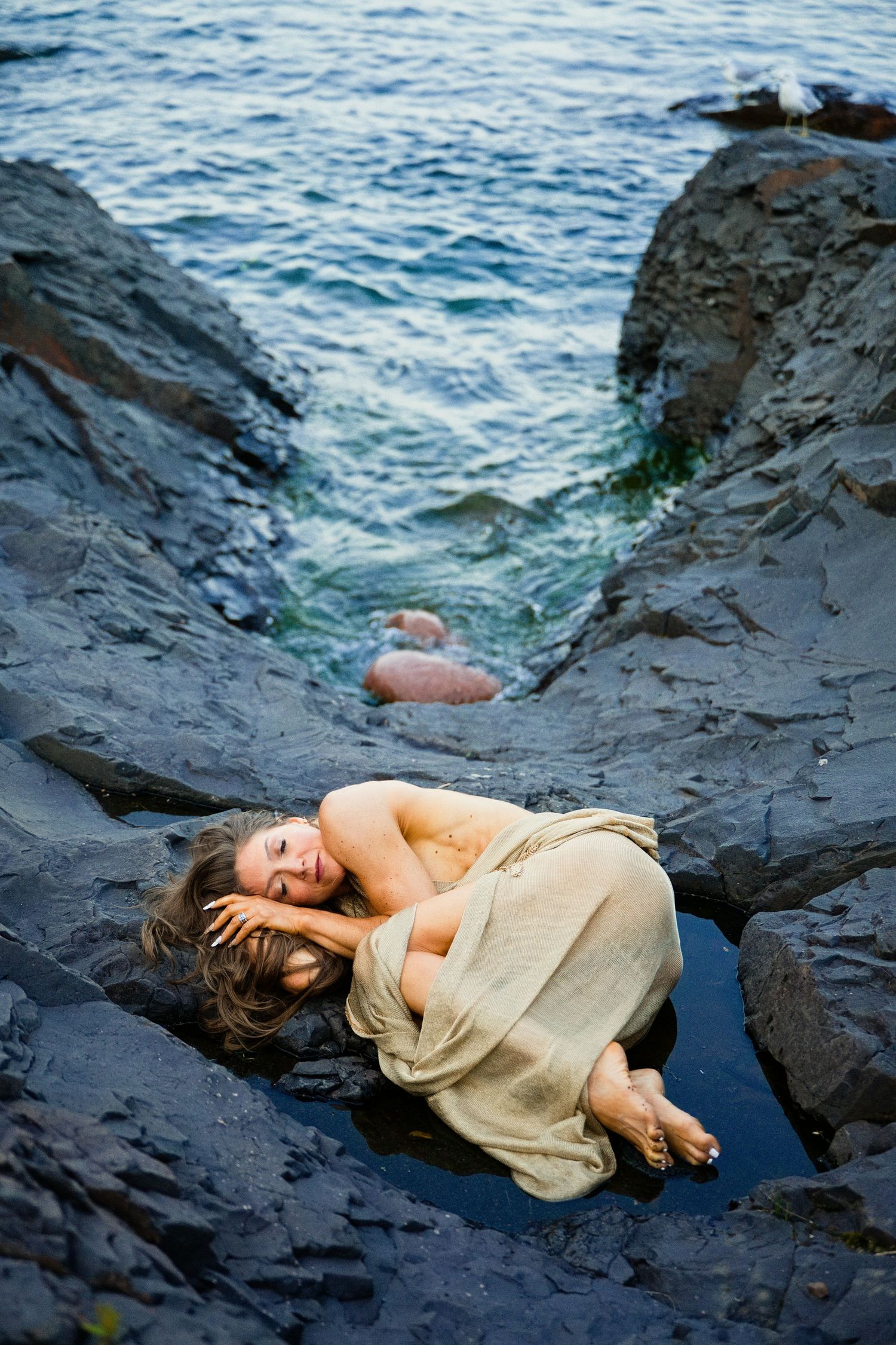 Woman in linen cloth lying on dark coastal rocks near ocean waves in a dramatic artistic pose.
