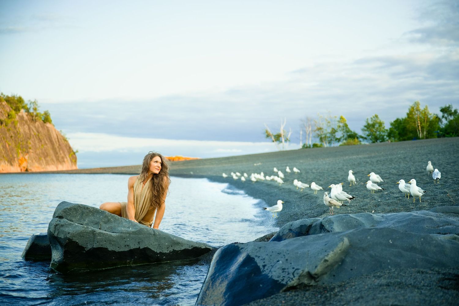 Woman with long hair sits on a rock by a calm shoreline with white seagulls nearby at golden hour.