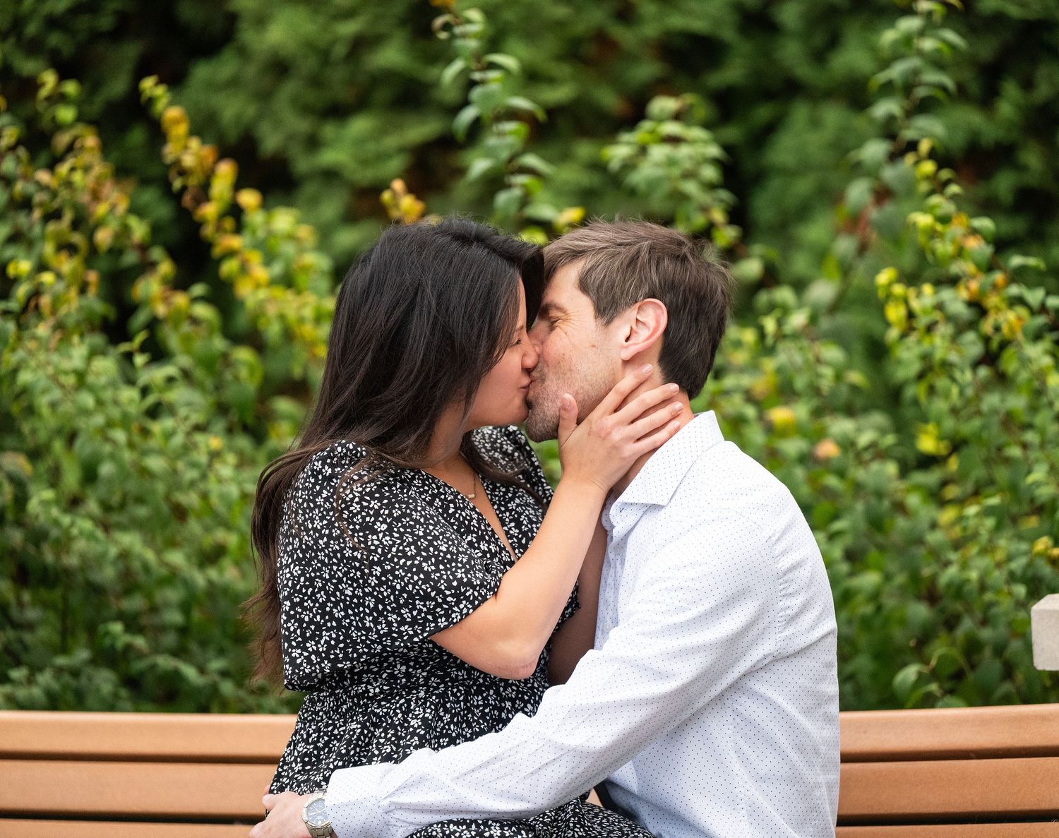 A couple shares a romantic kiss while sitting on a wooden bench in a lush garden setting at Lake Como. Engagement Sessions Minnesota.