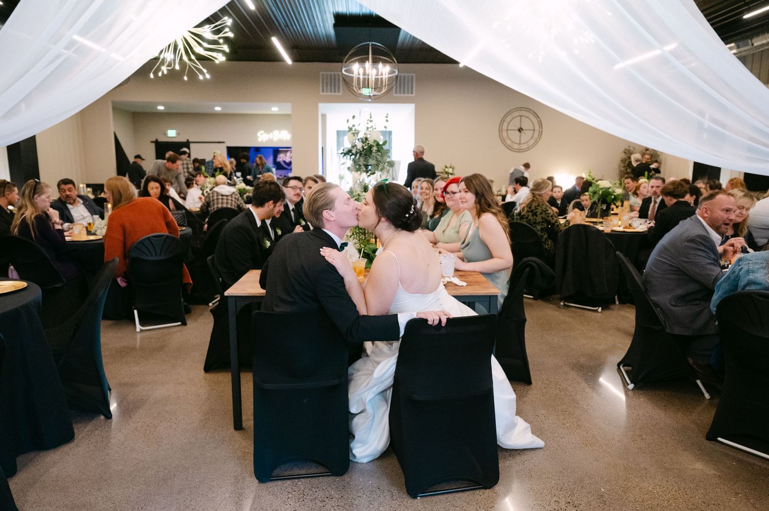 Wedding reception venue decorated with white drapes and black chair covers as guests mingle at round tables.
