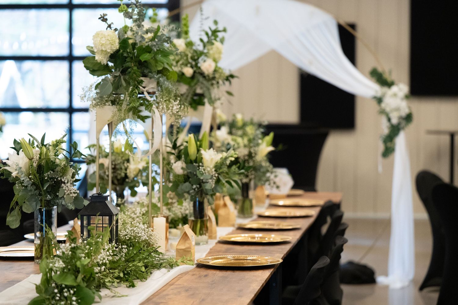Elegant wedding reception table with white floral centerpieces, greenery, and draped fabric decor in natural lighting.