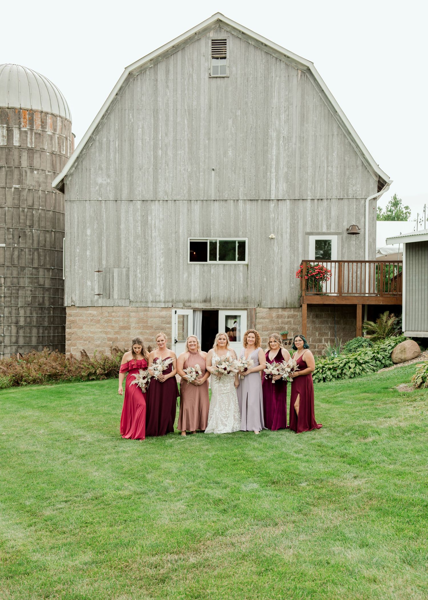 Wedding party members stand in front of a rustic white barn on a grassy lawn.
