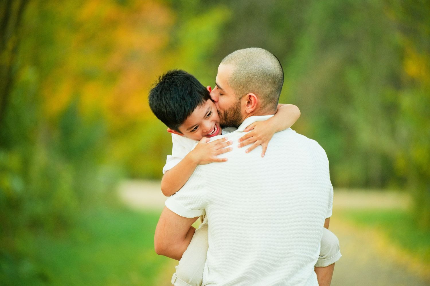 A father hugging his young son outdoors on a tree-lined path in autumn.