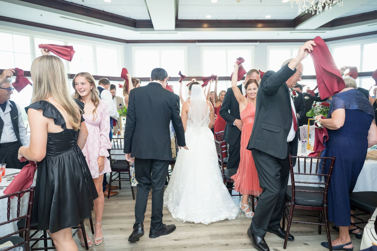 Wedding couple walks through guests waving red fabric at elegant indoor reception venue.