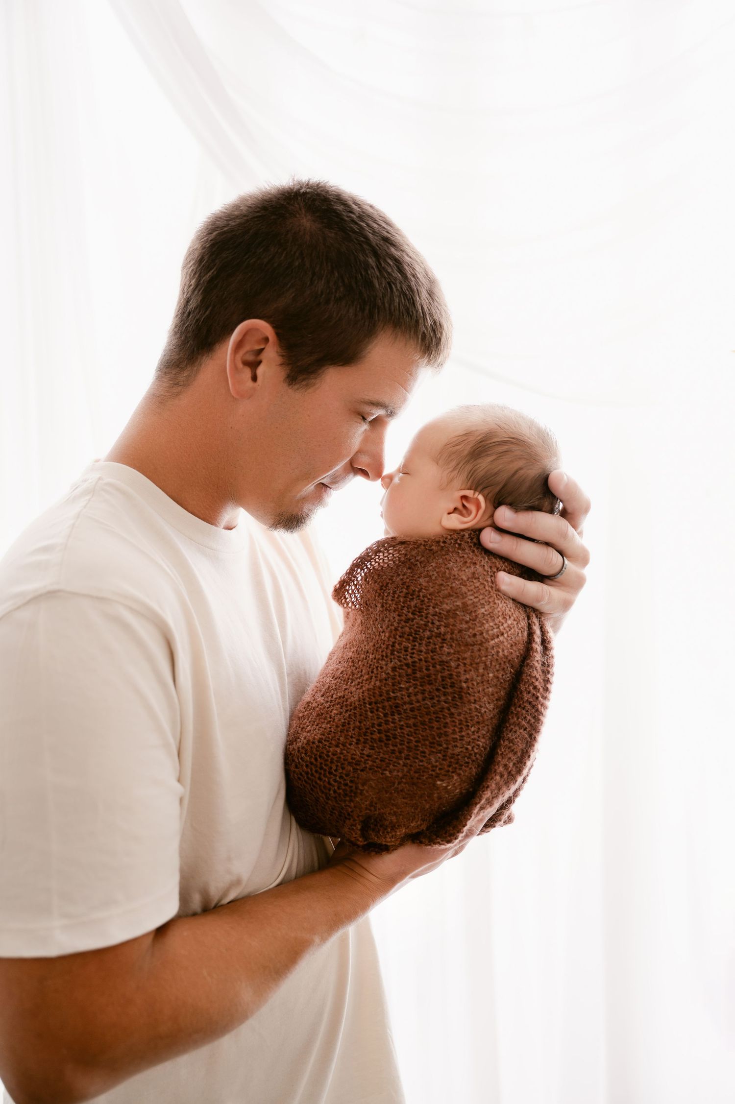 Tender moment between parent and newborn wrapped in brown knit blanket against white backdrop.