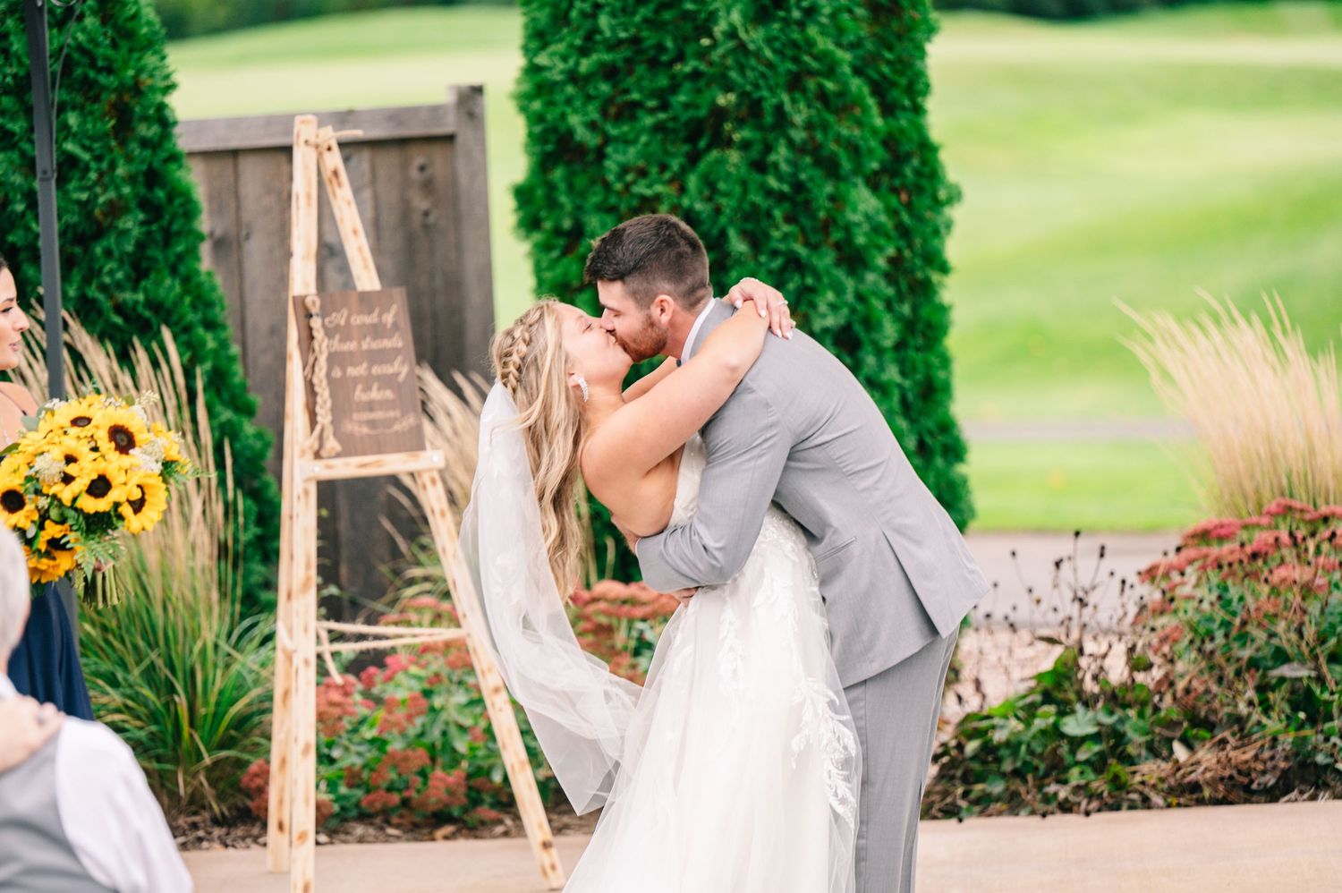 Central MN Wedding Photos - Bride and groom share first kiss dip at outdoor wedding ceremony near sunflower decorations.