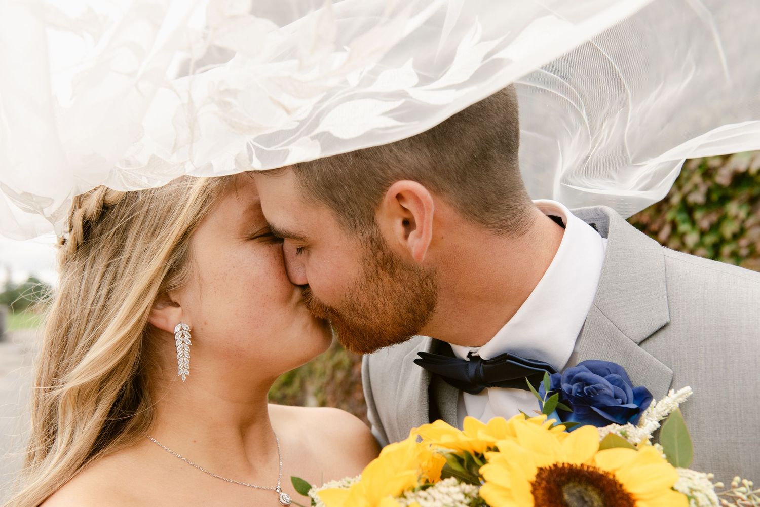 Central MN Wedding Photos | Bride and groom kissing under flowing veil, bride holding sunflower and blue rose bouquet.
