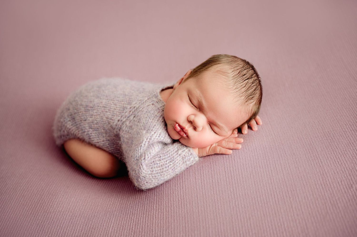 Sleeping newborn baby in lavender knit romper posed on pink blanket for professional newborn photography.