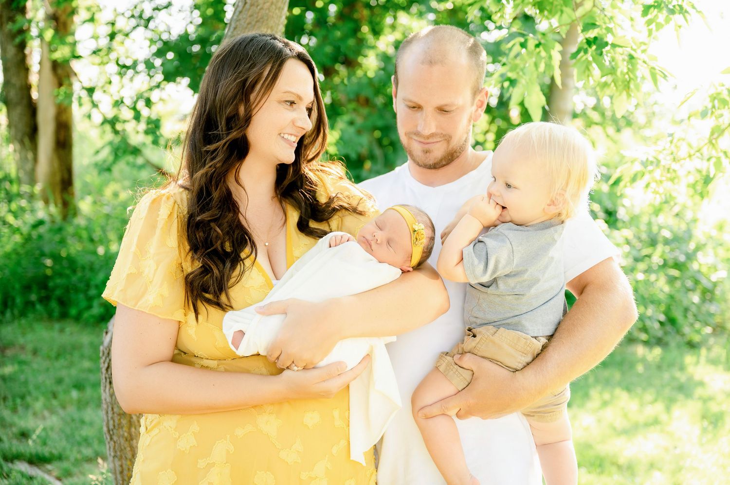 St Cloud newborn Photos of A young family poses outdoors, mother in yellow holds newborn, father holds toddler, surrounded by green trees.