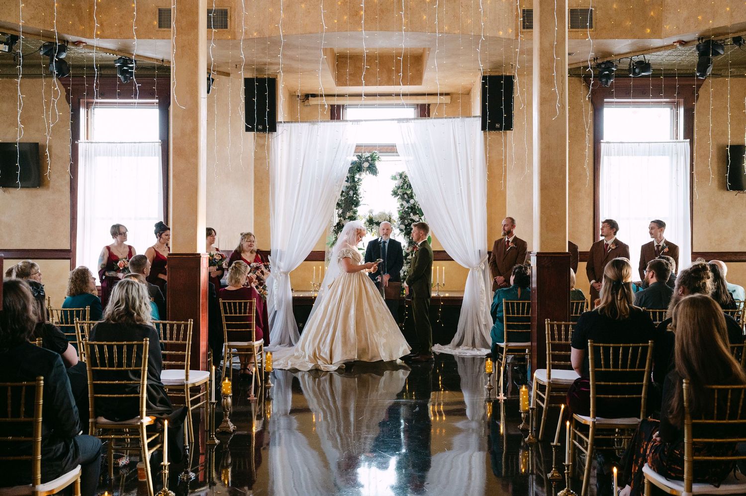 Central MN Wedding Photos | Bride and groom share first kiss at indoor wedding ceremony with fairy lights.