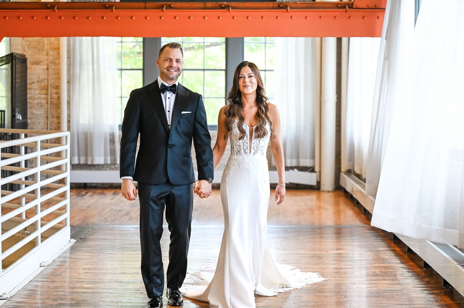 Central MN Wedding Photos | Bride and groom holding hands in elegant wedding attire inside a rustic industrial venue with hardwood floors.